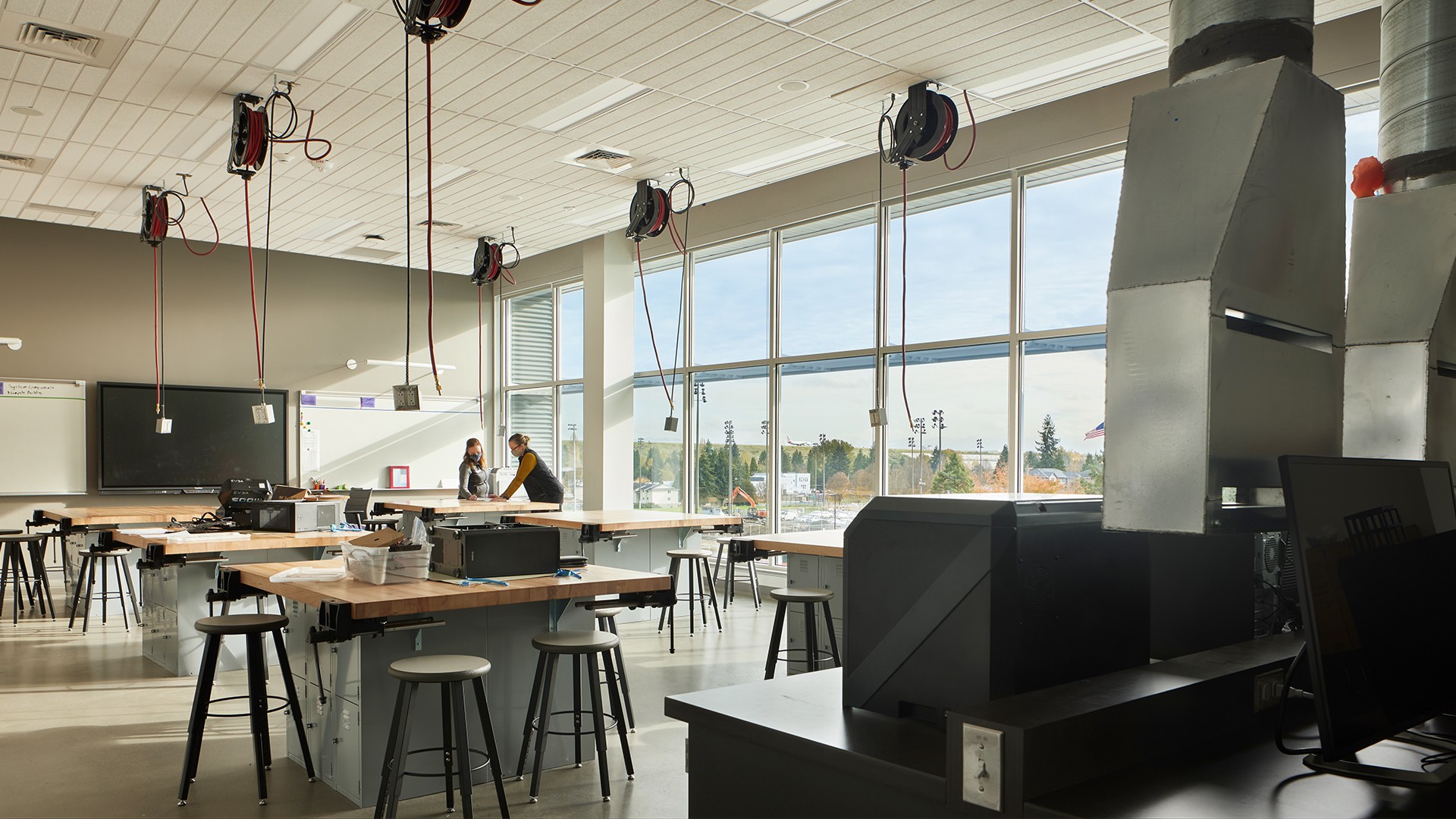 Modern classroom with large windows, workbenches, stools, and two people talking.