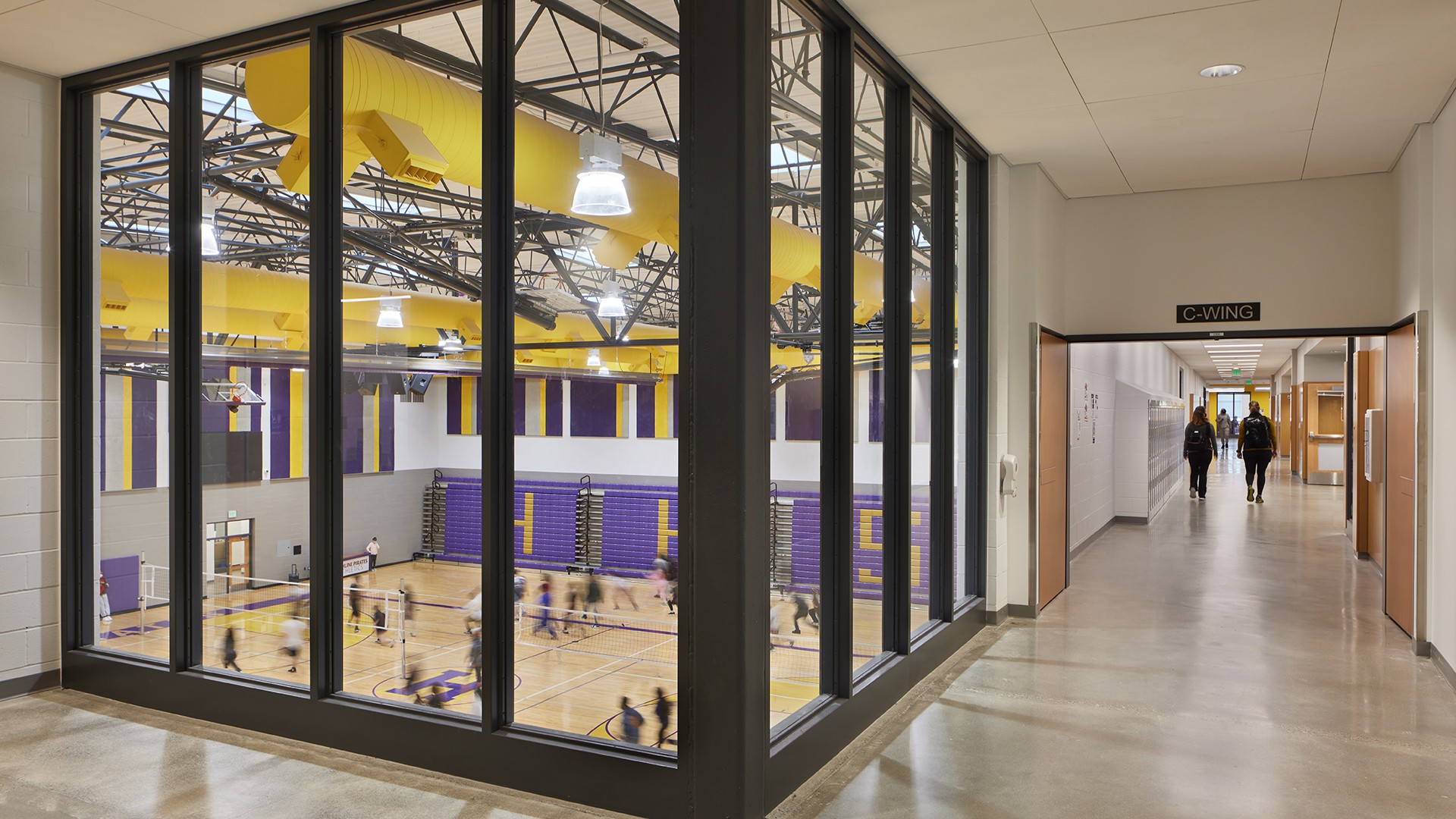 Modern school gymnasium with students playing basketball, viewed from a hallway.