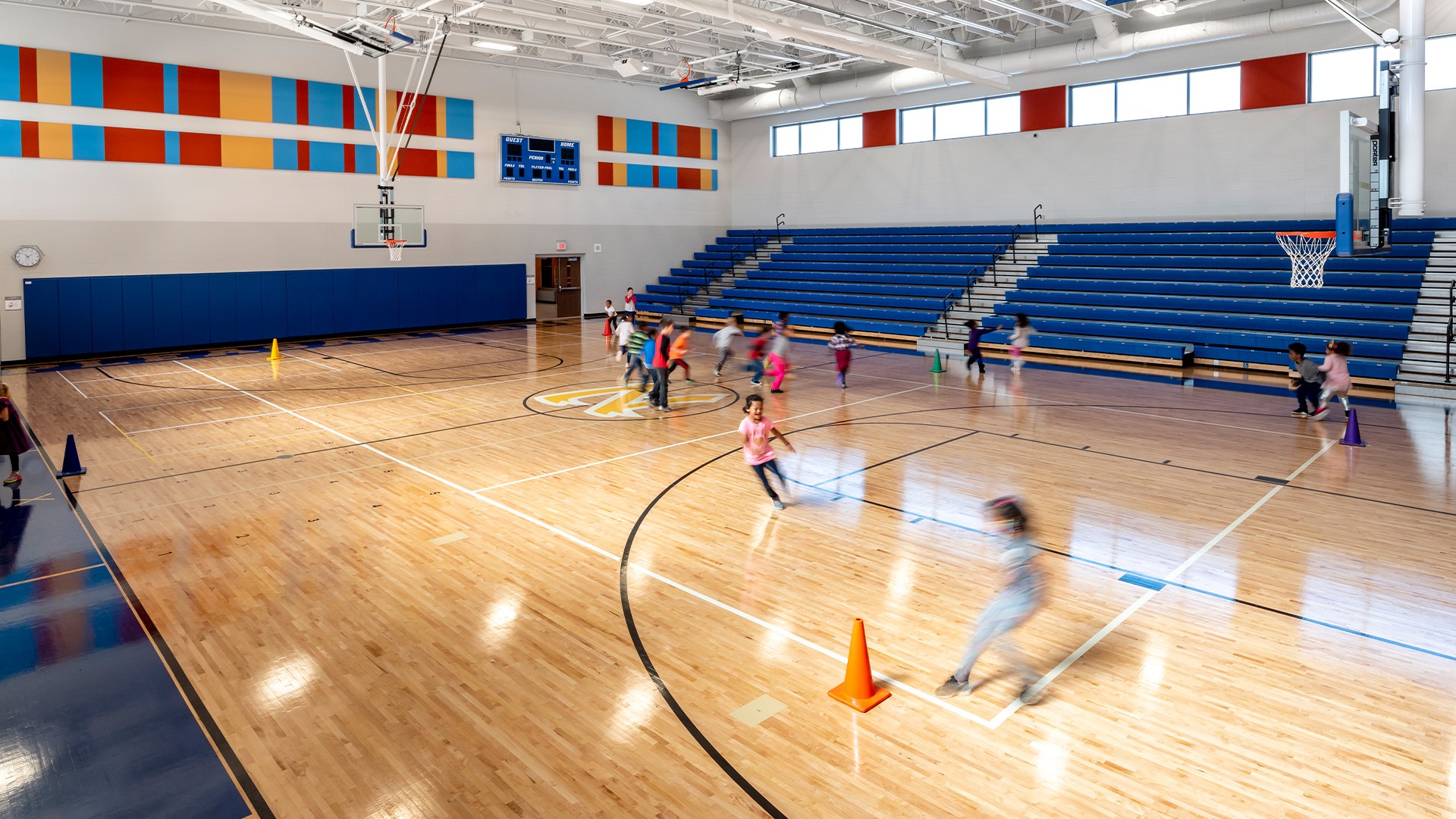 Children playing and running in a bright, spacious indoor school gymnasium with bleachers.