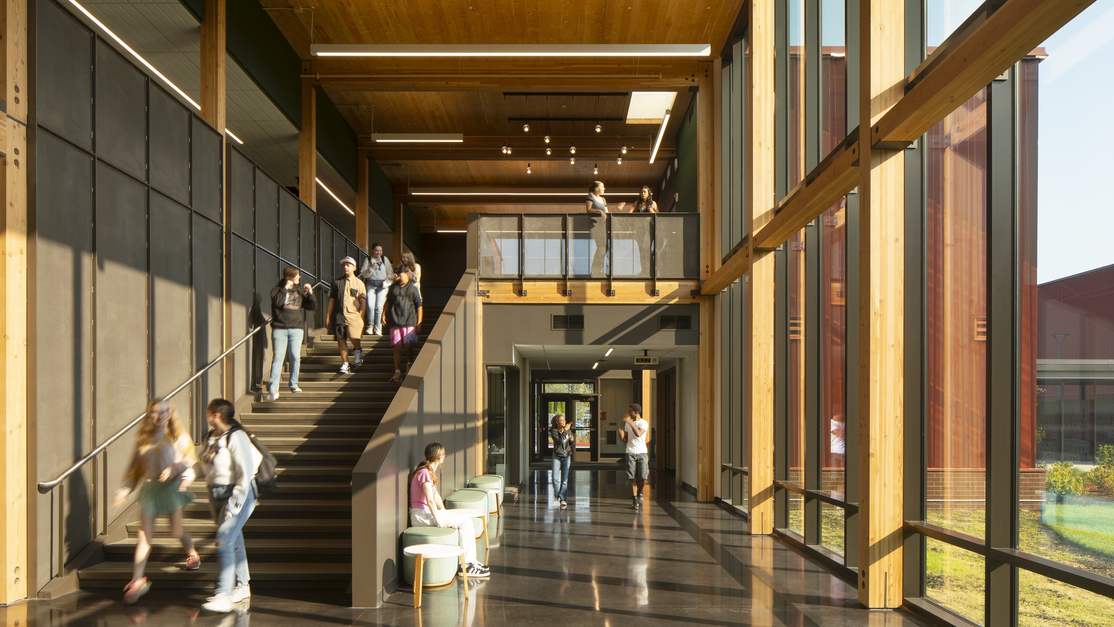 Modern school hallway with large windows, wooden beams, and students on stairs and benches.