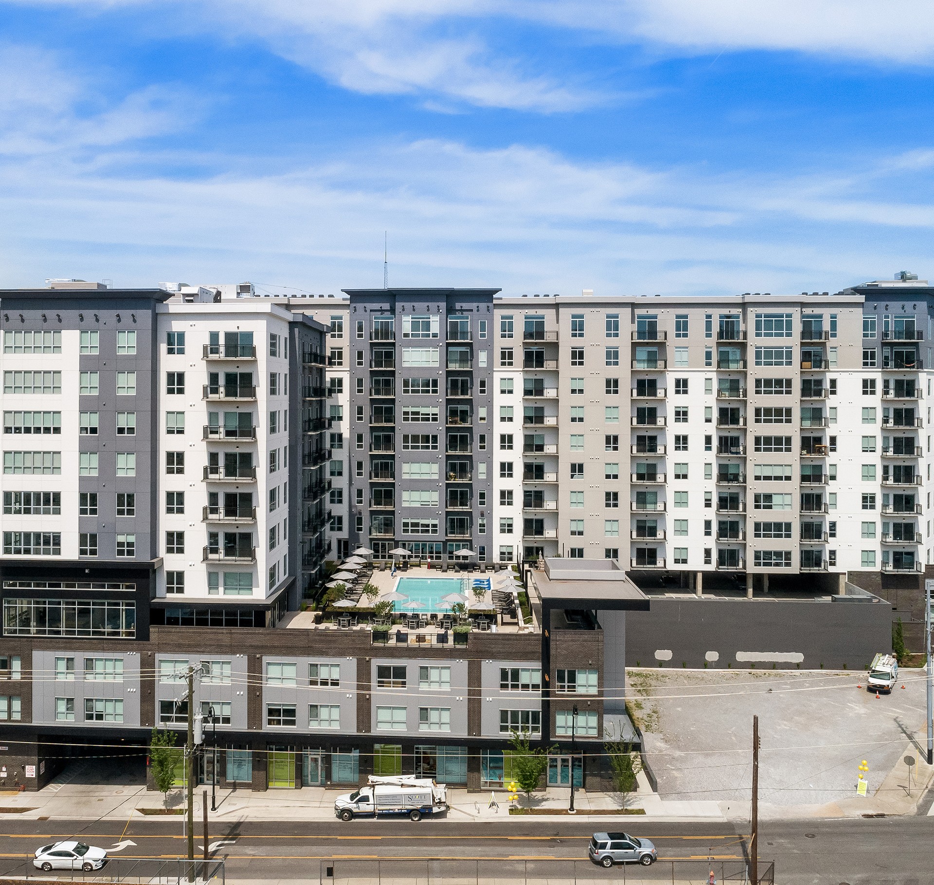 Modern Apartment Complex with Rooftop Pool and Parking Area Modern multi-story apartment building with rooftop pool under a blue sky.