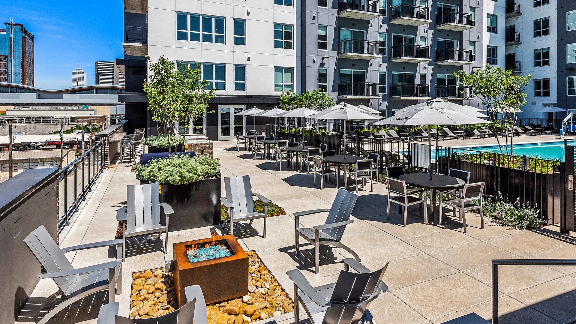Modern outdoor patio with chairs, fire pit, umbrellas, and a swimming pool in a city.