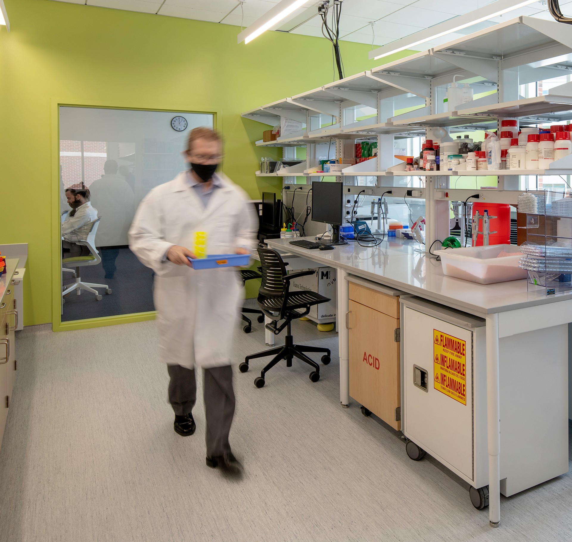 Modern Scientific Laboratory with Safety Equipment and Supplies A scientist in a lab coat carries equipment through a modern laboratory with shelves and workstations.