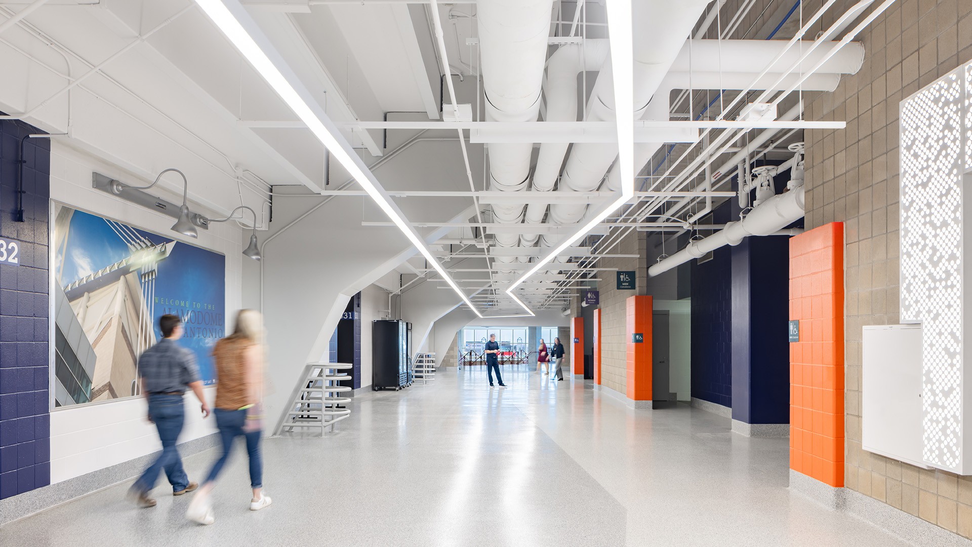 Modern bright hallway with white ceiling pipes, orange pillars, and people walking.