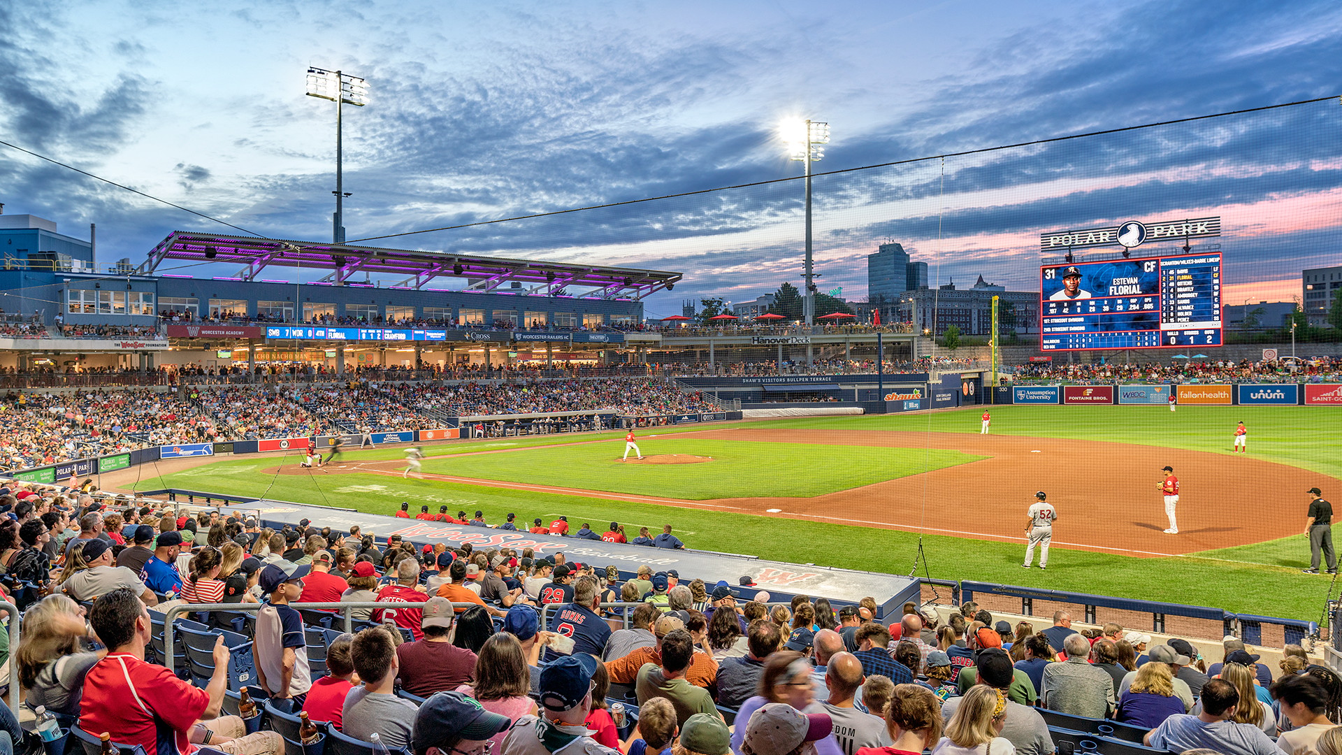 Crowded Polar Park baseball stadium at sunset with players on the field and scoreboard.