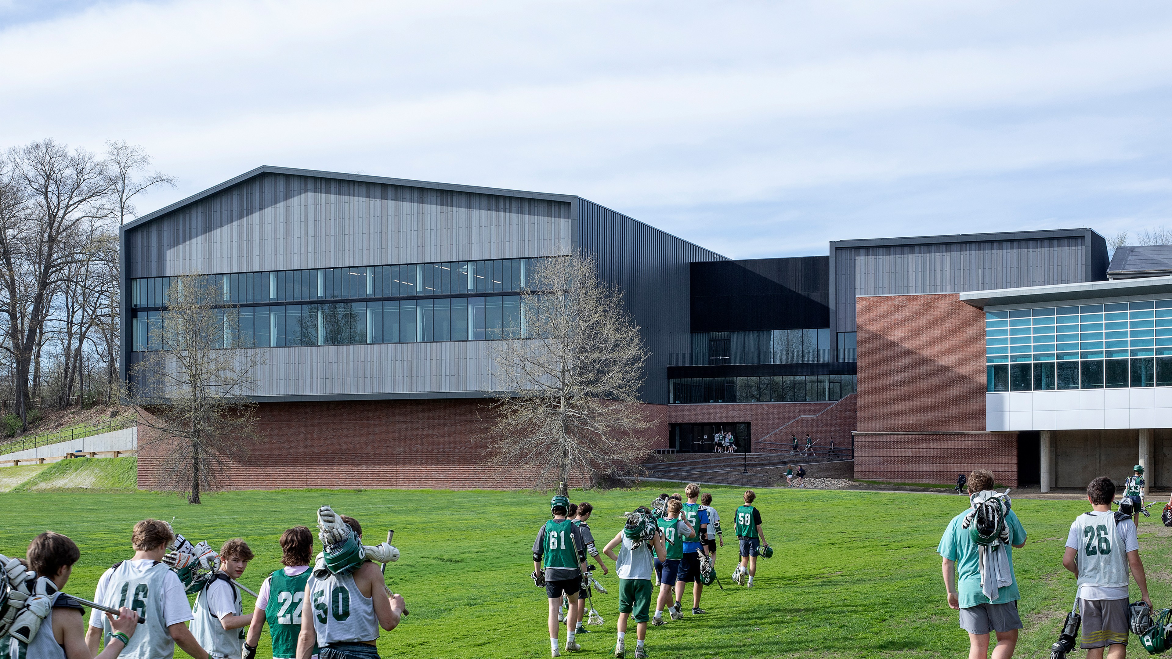 Students carrying lacrosse gear walk across a green field toward a modern school building.