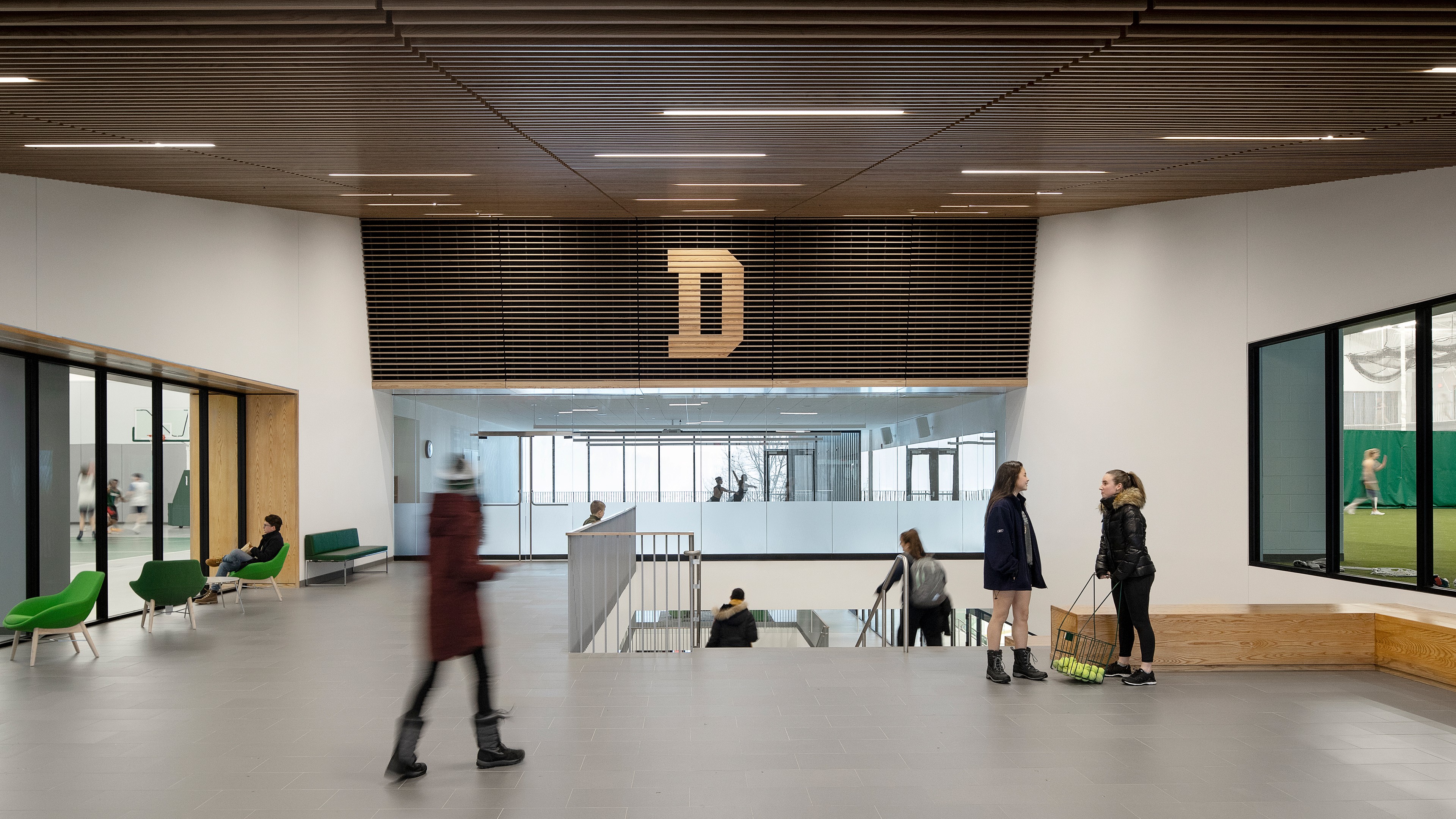 Modern indoor sports facility lobby with people and green chairs under wooden ceiling.