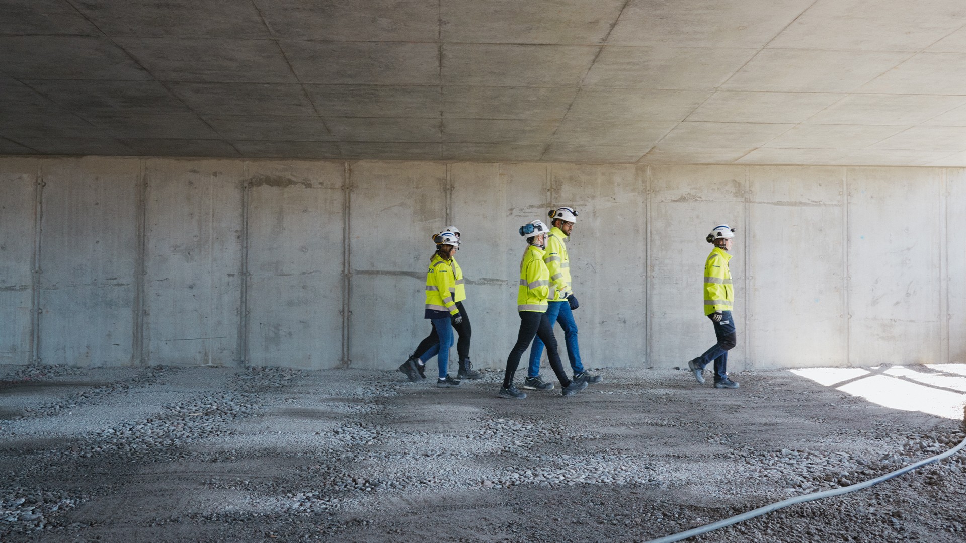 Construction workers in safety gear walk under a concrete structure at a building site.