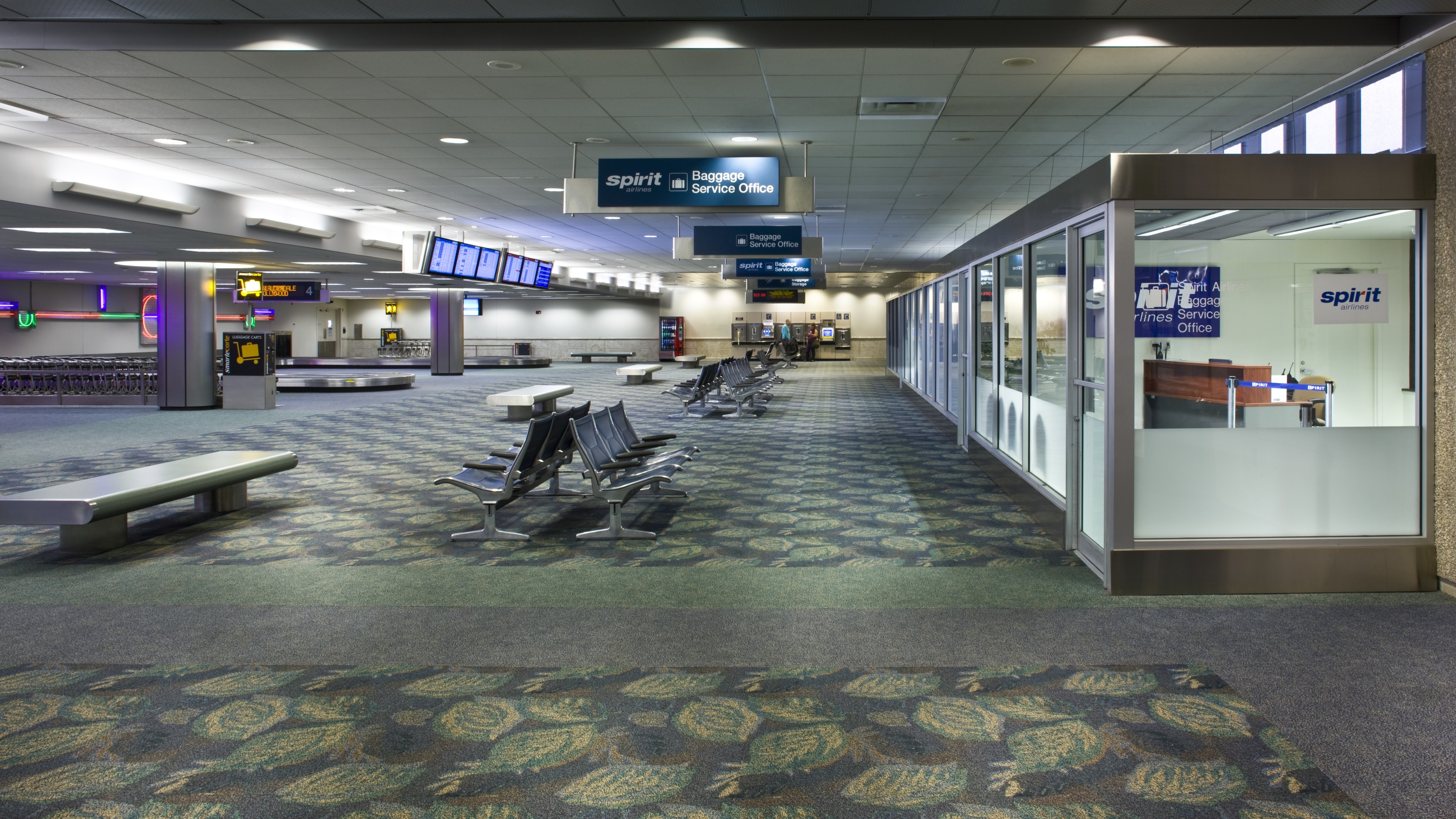 Empty airport baggage claim area with Spirit Airlines service office and seating.