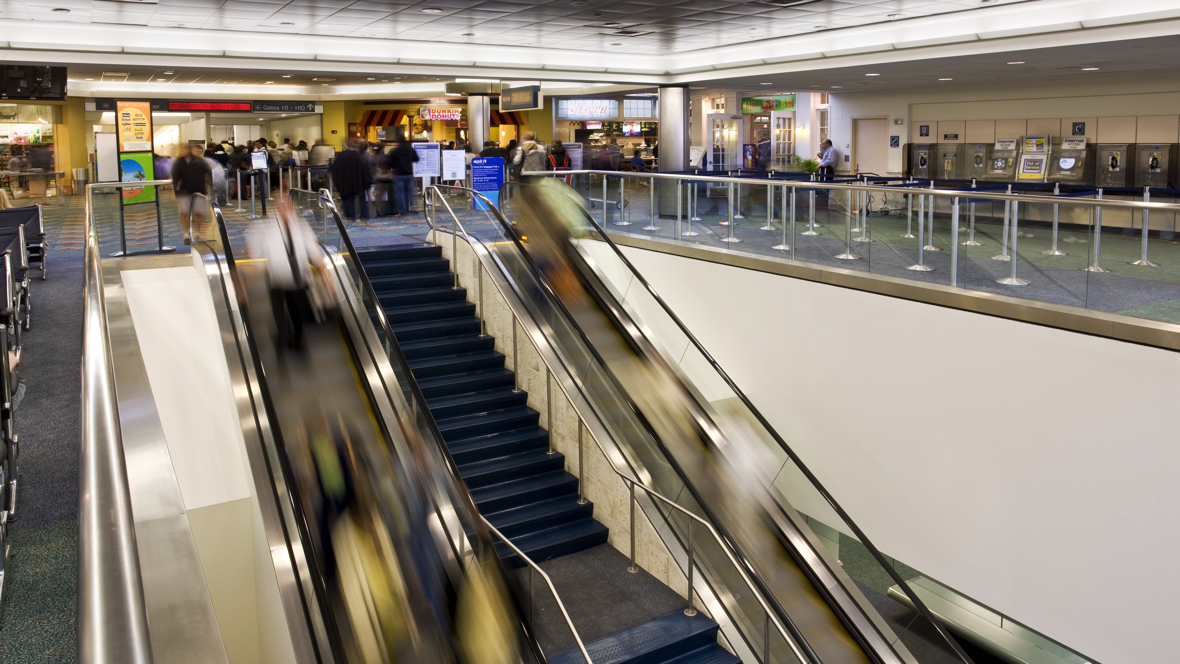 Busy airport terminal with escalators, stairs, and travelers moving between levels.