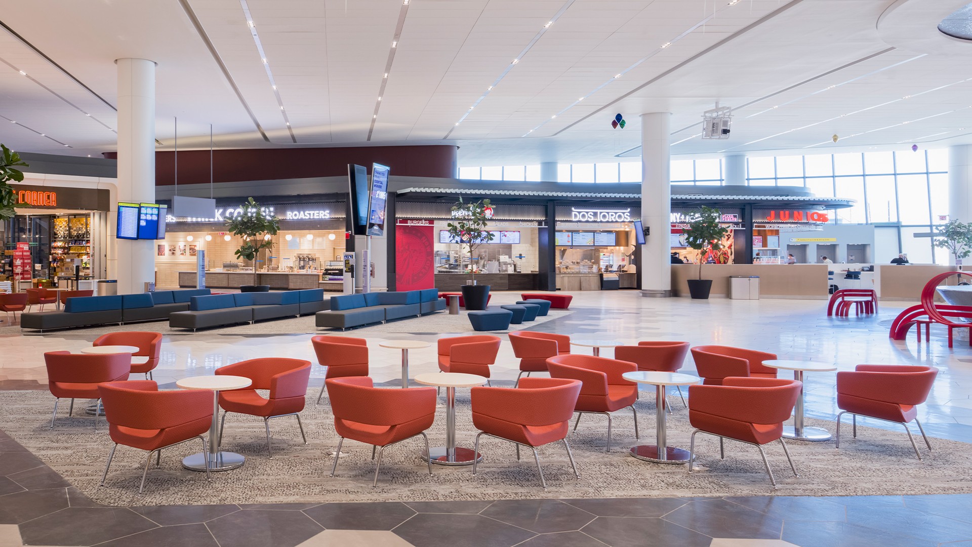 Modern airport lounge area with red chairs, tables, and shops in the background.