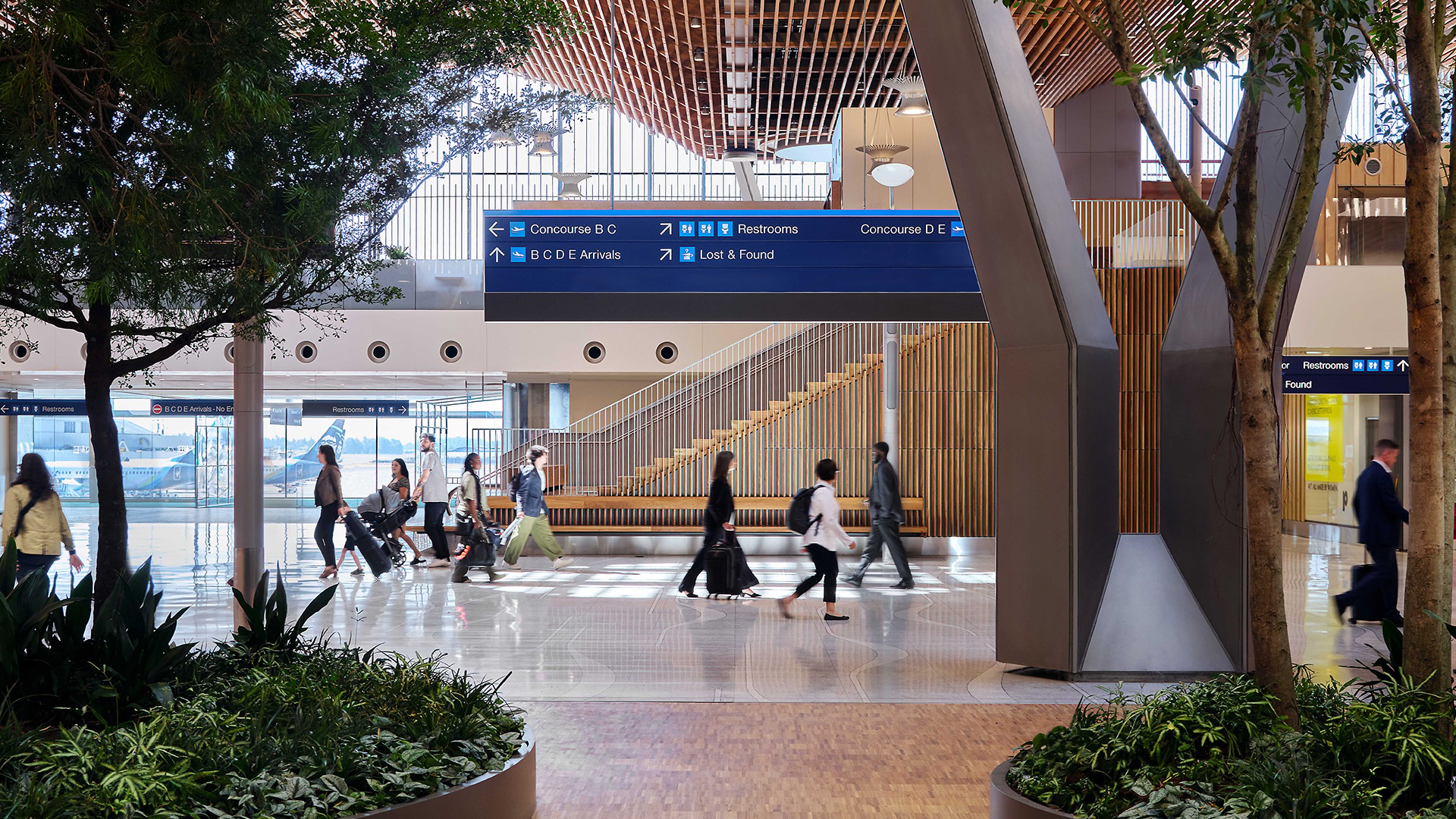Busy airport terminal with travelers walking and directional signs overhead.