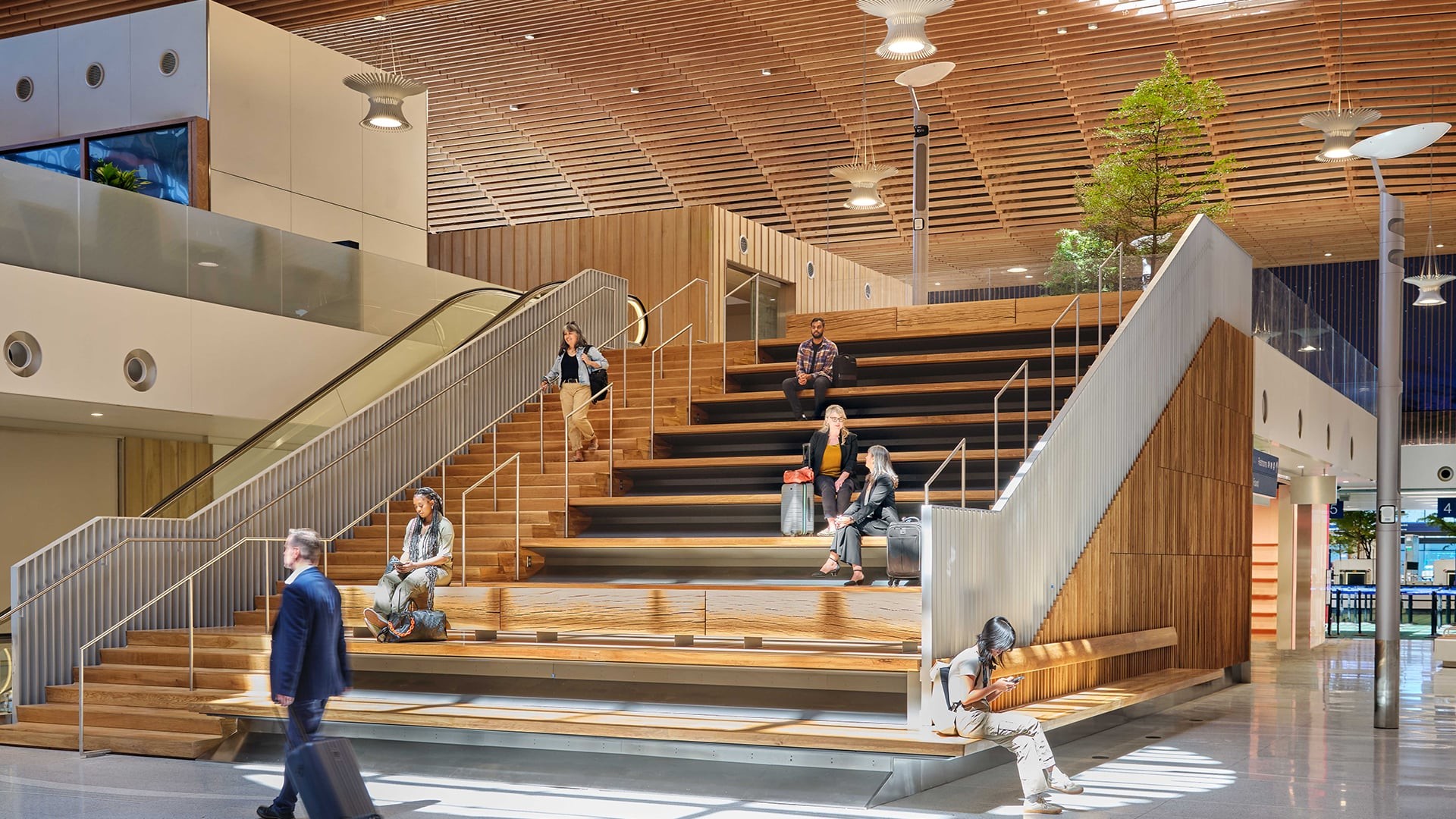 Modern airport lounge with wooden seating steps, travelers, and natural lighting.