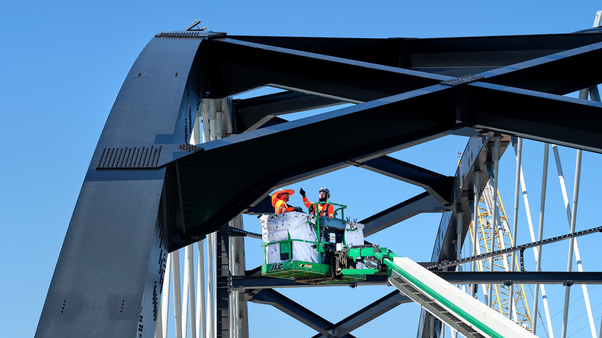 Workers Inspecting Steel Bridge Structure Using Aerial Lift Workers in safety gear inspecting a steel bridge structure using a green lift platform.