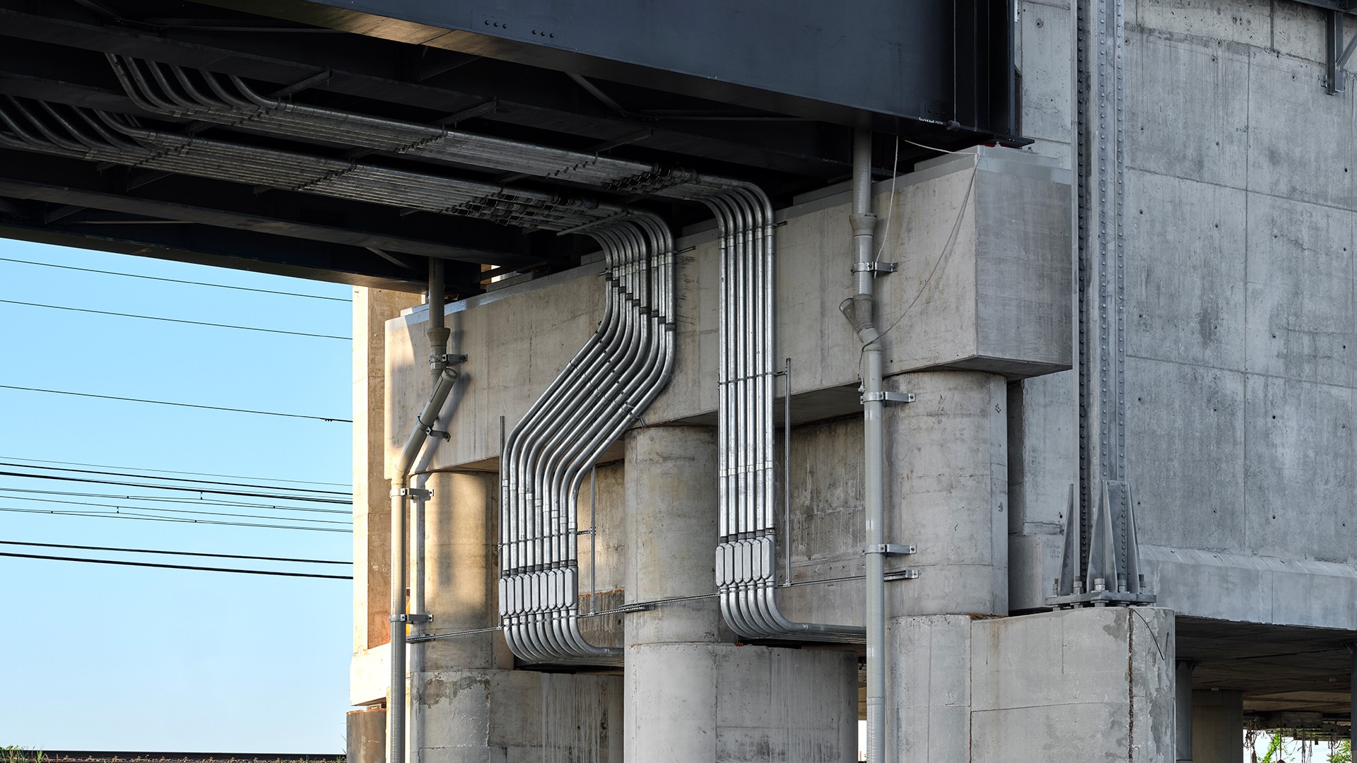 Industrial concrete structure with metal pipes under bridge Concrete bridge pillars with metal electrical conduits and wiring under a clear sky.