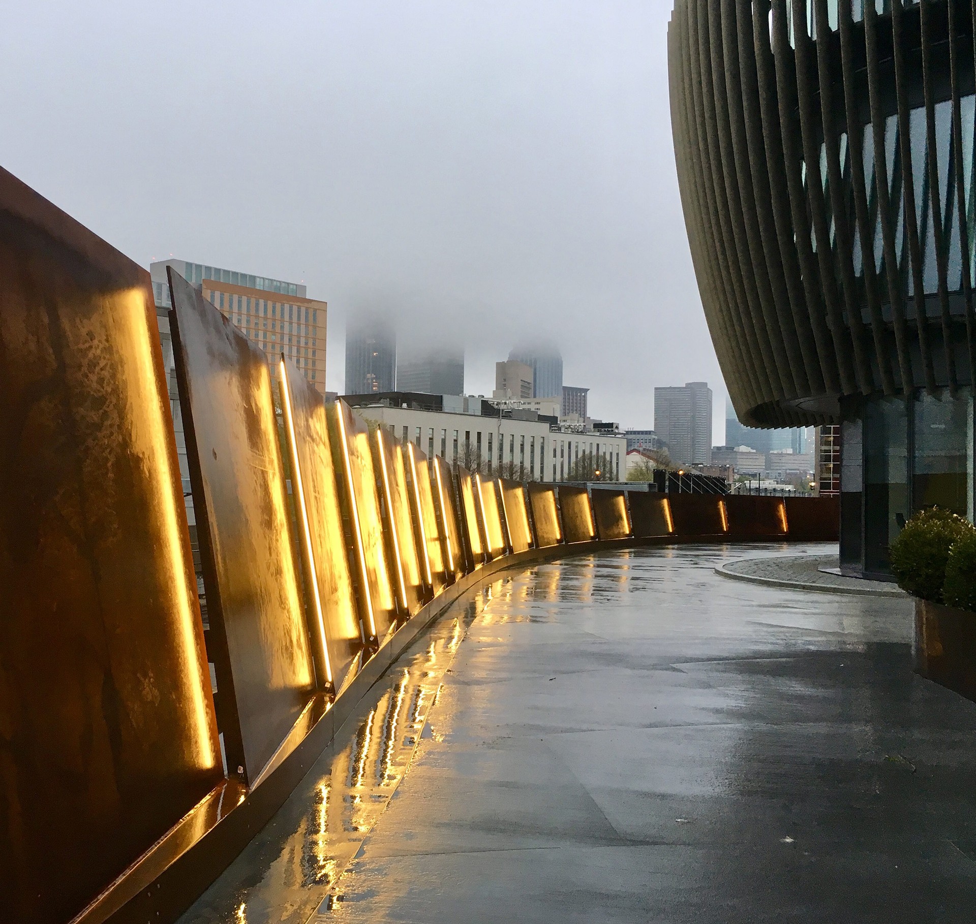 Curved illuminated metal panels along a wet urban walkway with foggy city skyline.