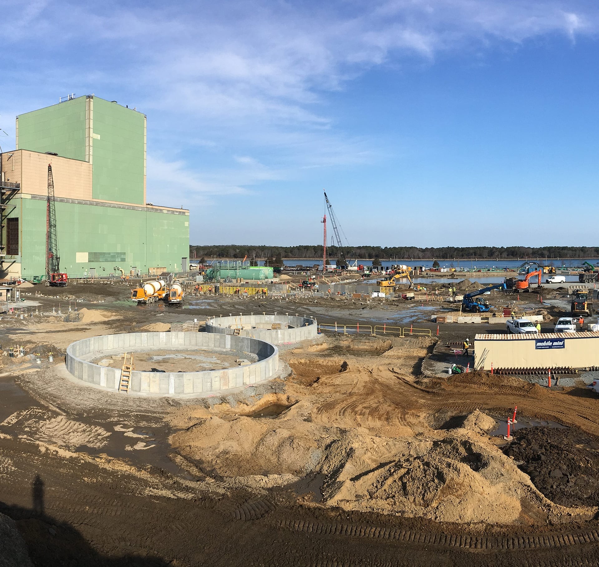 Industrial construction site with large green building and cranes Construction site with large circular concrete foundations and heavy machinery under a blue sky.