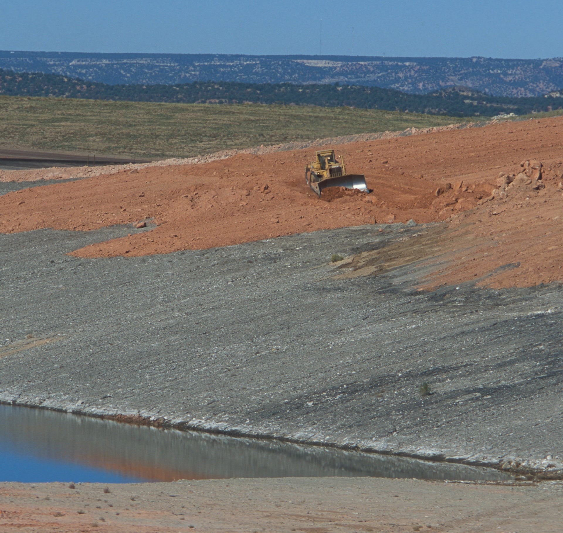 Bulldozer grading red soil on a rocky slope near a water body in a rural landscape.