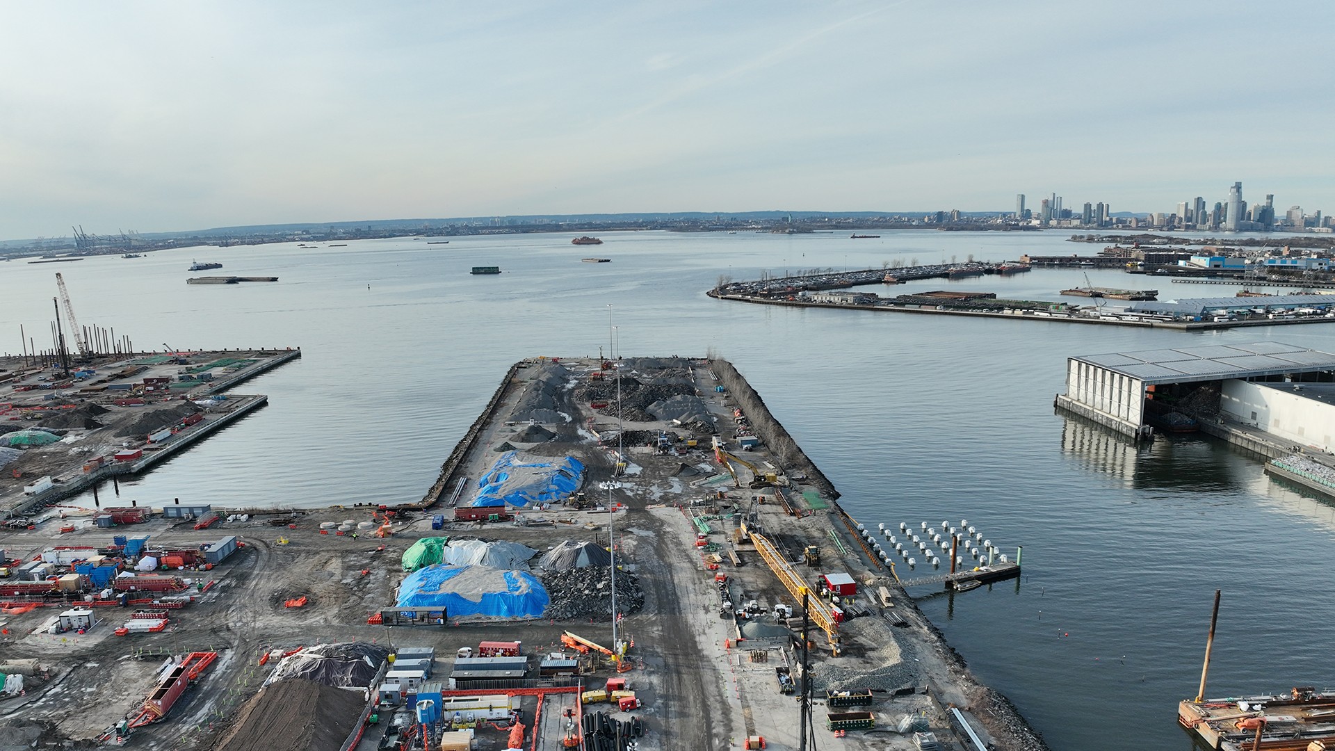Aerial view of a coastal construction site with equipment and city skyline in the distance.