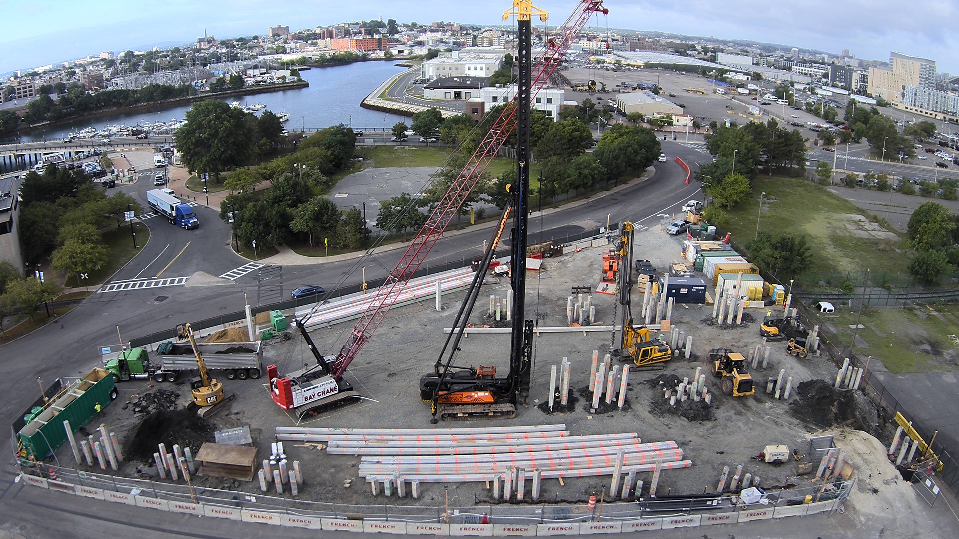 Construction site with cranes and concrete piles near a waterfront urban area.