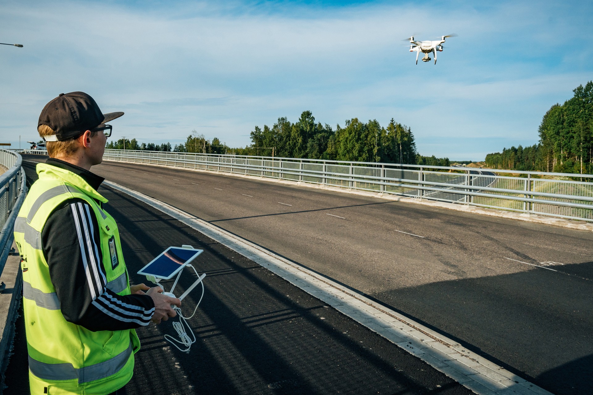 A person operates a drone over a bridge, using a remote control and tablet, wearing a safety vest.