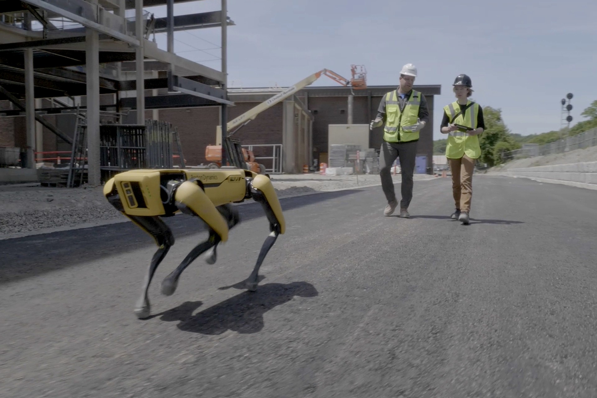 Robotic Dog on Construction Site with Engineers Observing Two construction workers follow a yellow robotic dog on a paved site near a building structure.