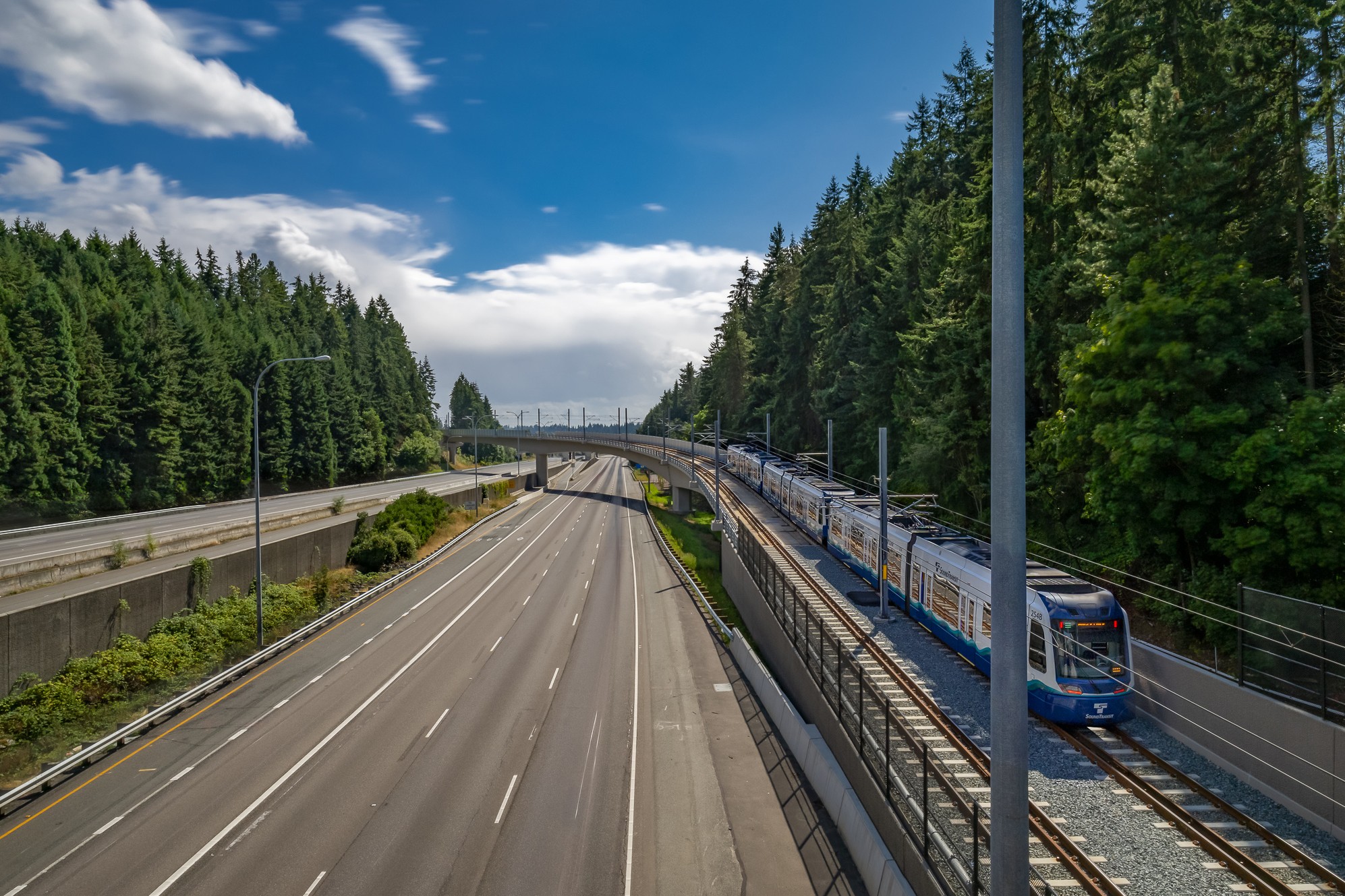 Modern light rail train running beside empty highway in forest Light rail train traveling on tracks beside an empty highway surrounded by green trees.