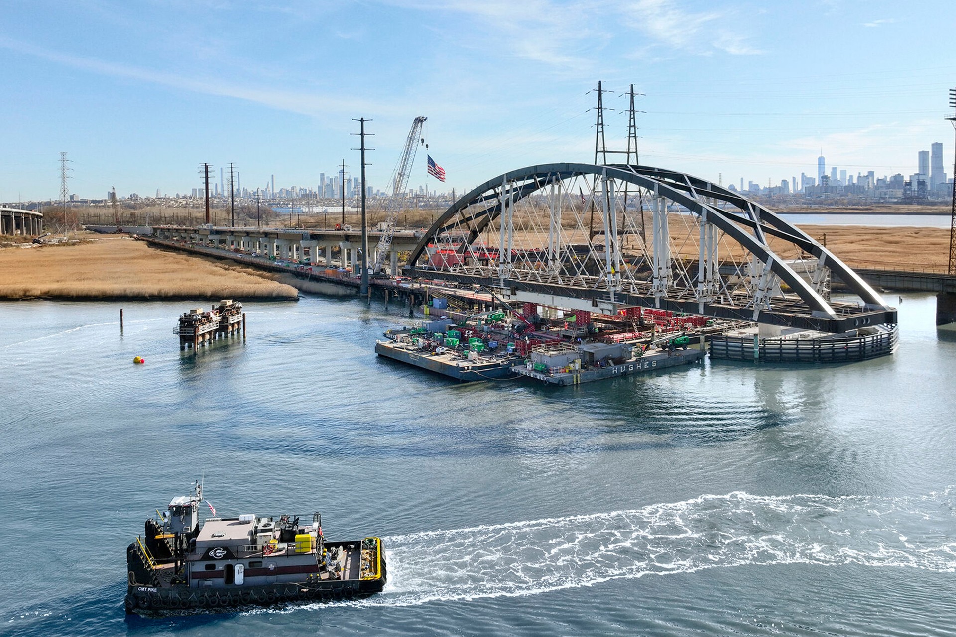 Tugboat passing under an arched bridge with construction barge and city skyline in background.
