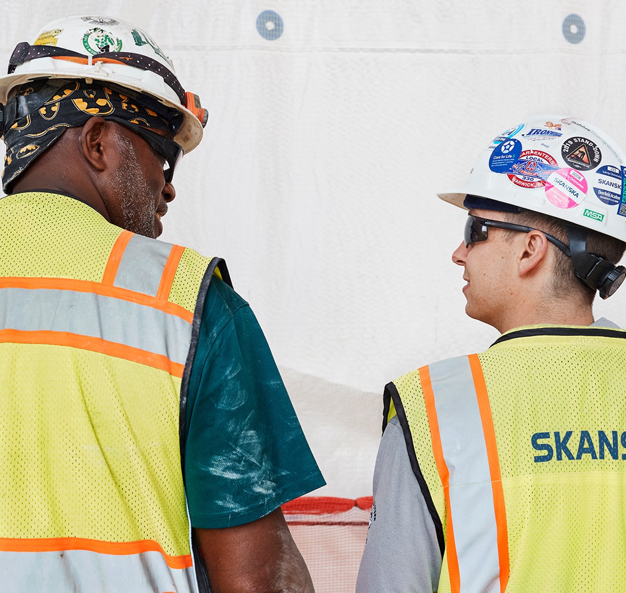Construction Workers in Safety Gear at Skanska Site Two construction workers wearing safety vests and hard hats with various stickers on a job site.