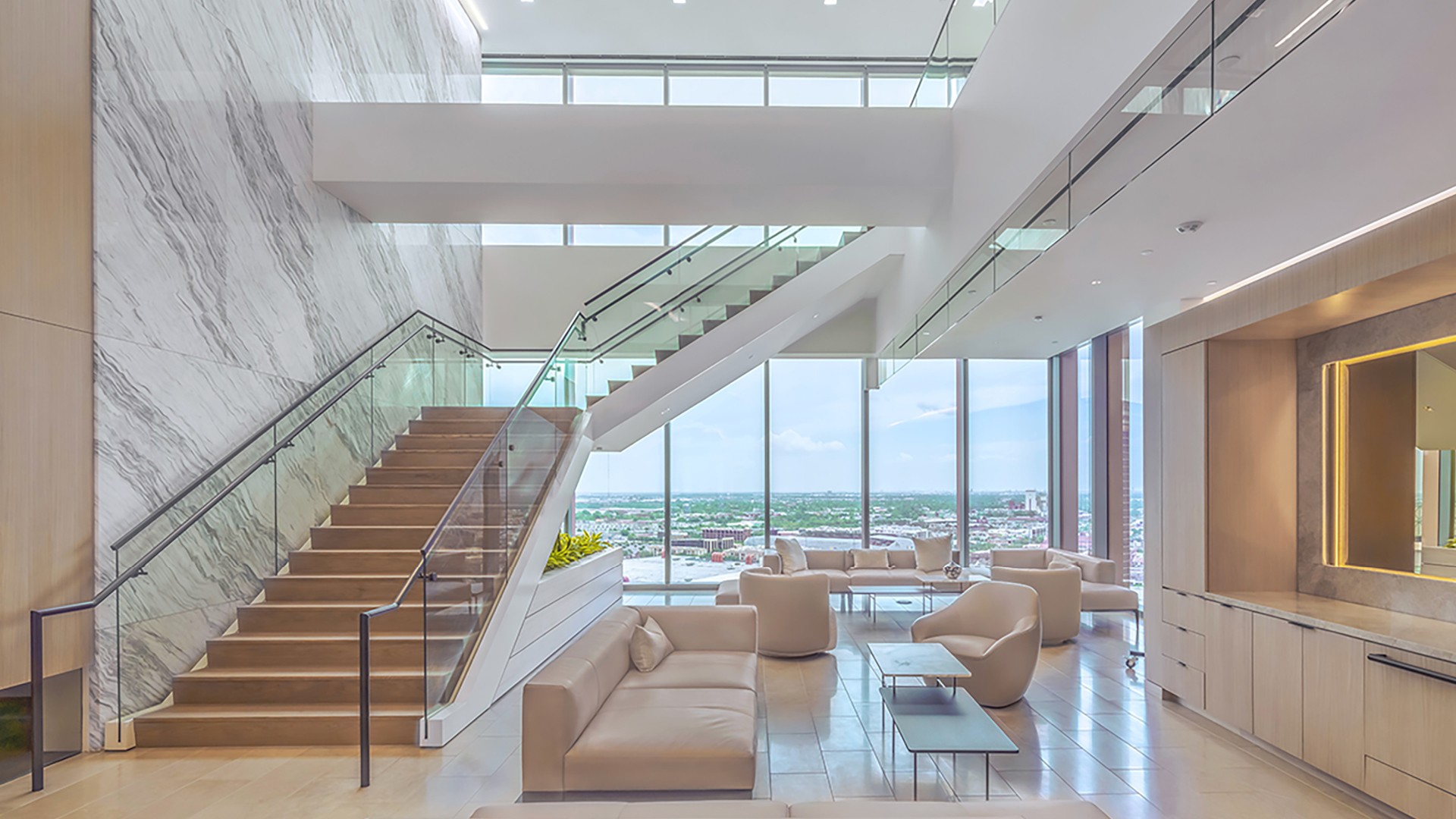 Modern office lounge with glass staircase, beige seating, and large city-view windows.