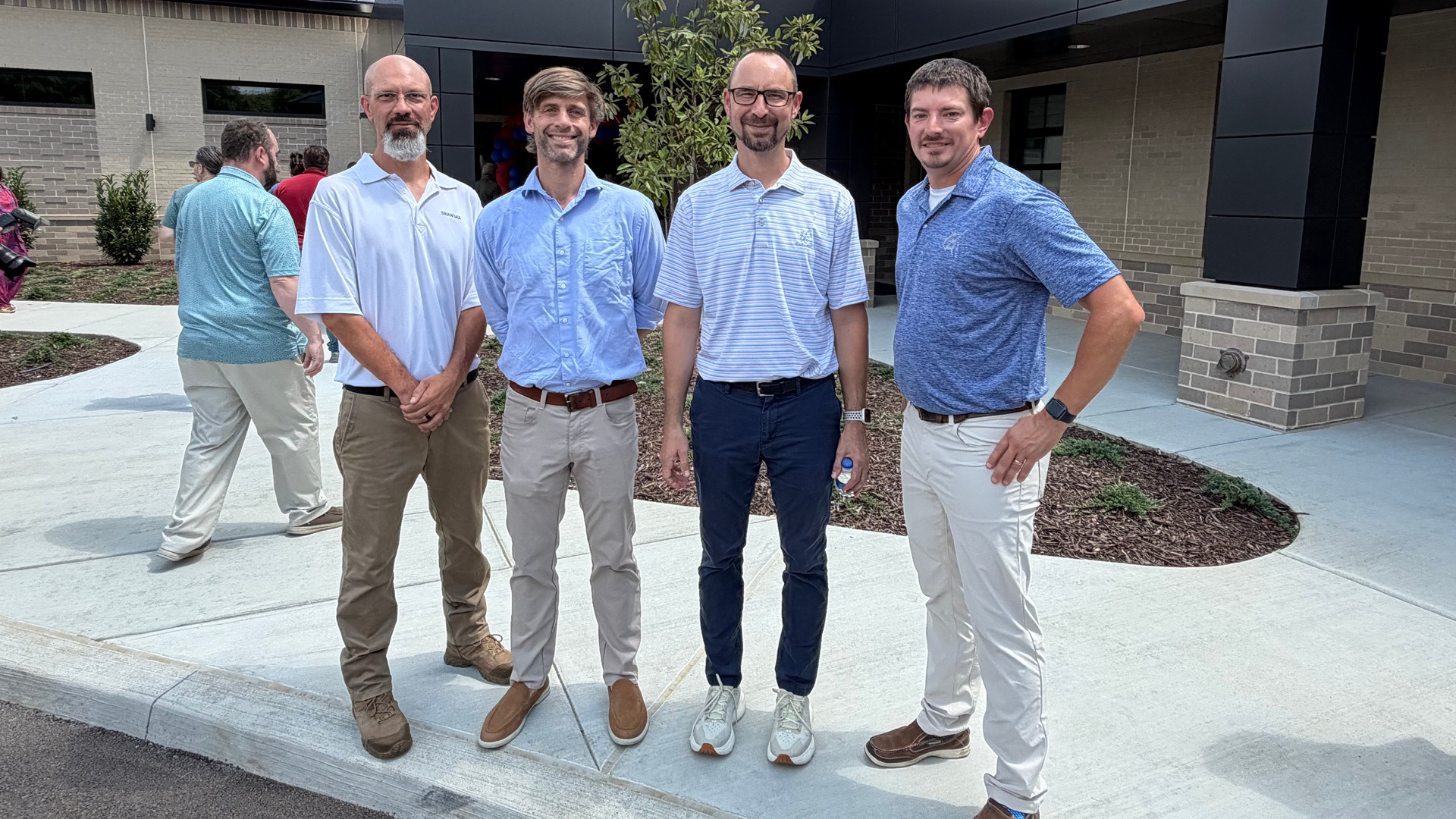 Four casually dressed men standing outdoors on a sidewalk near a modern building entrance.