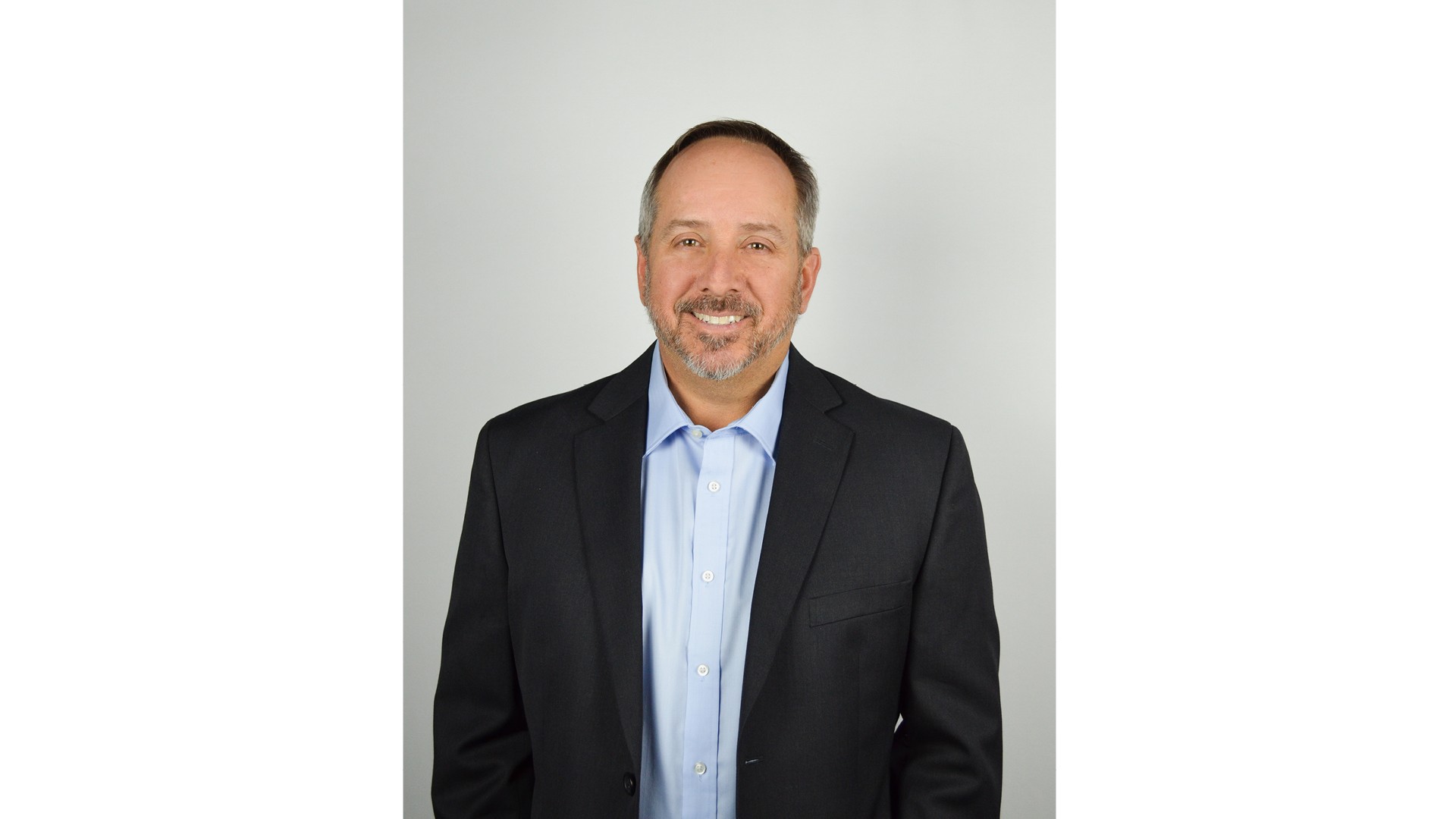 Professional middle-aged man in a dark blazer and light blue shirt smiling on gray background.
