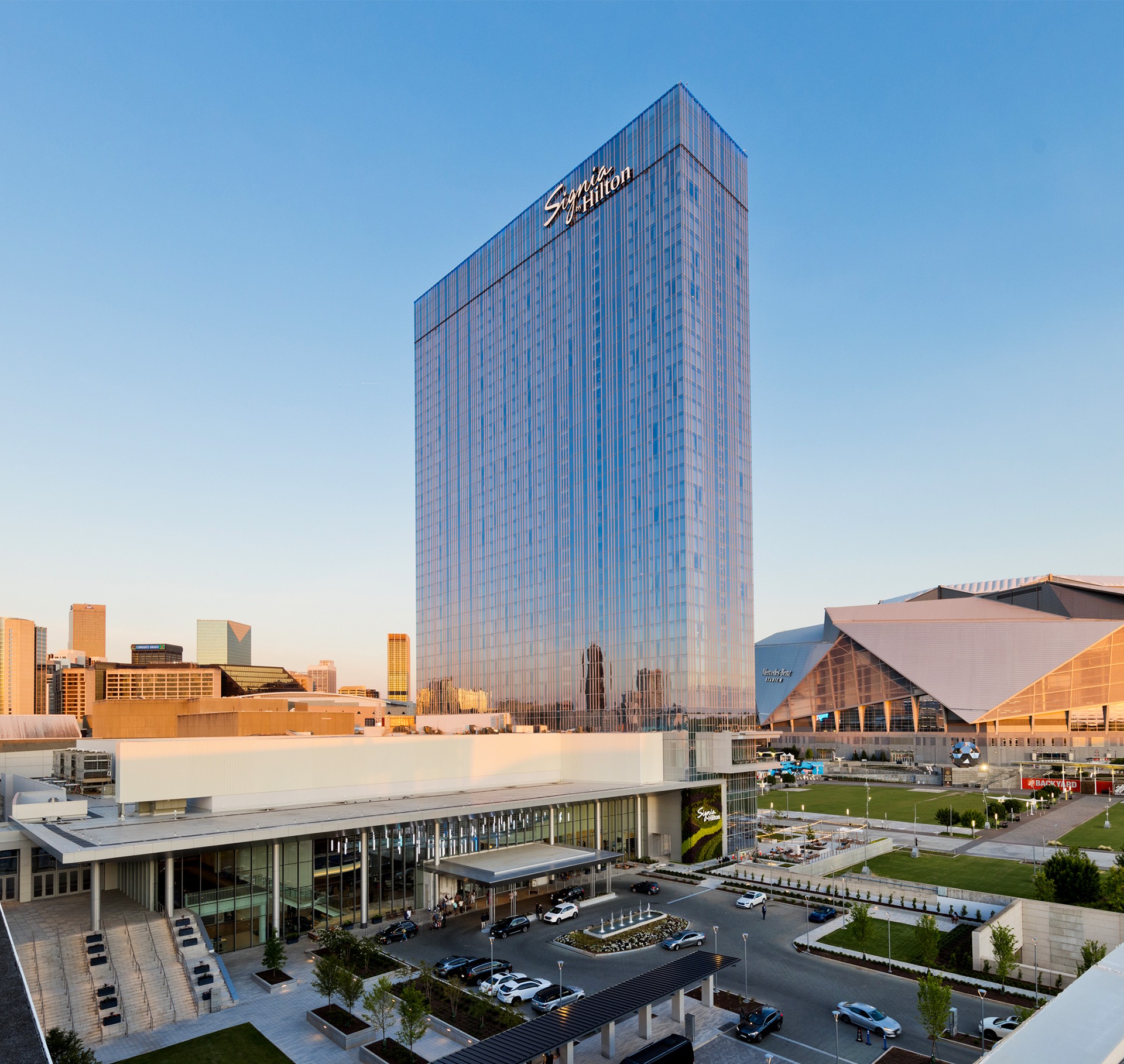 Modern skyscraper hotel with glass facade next to a large stadium in an urban setting at sunset.