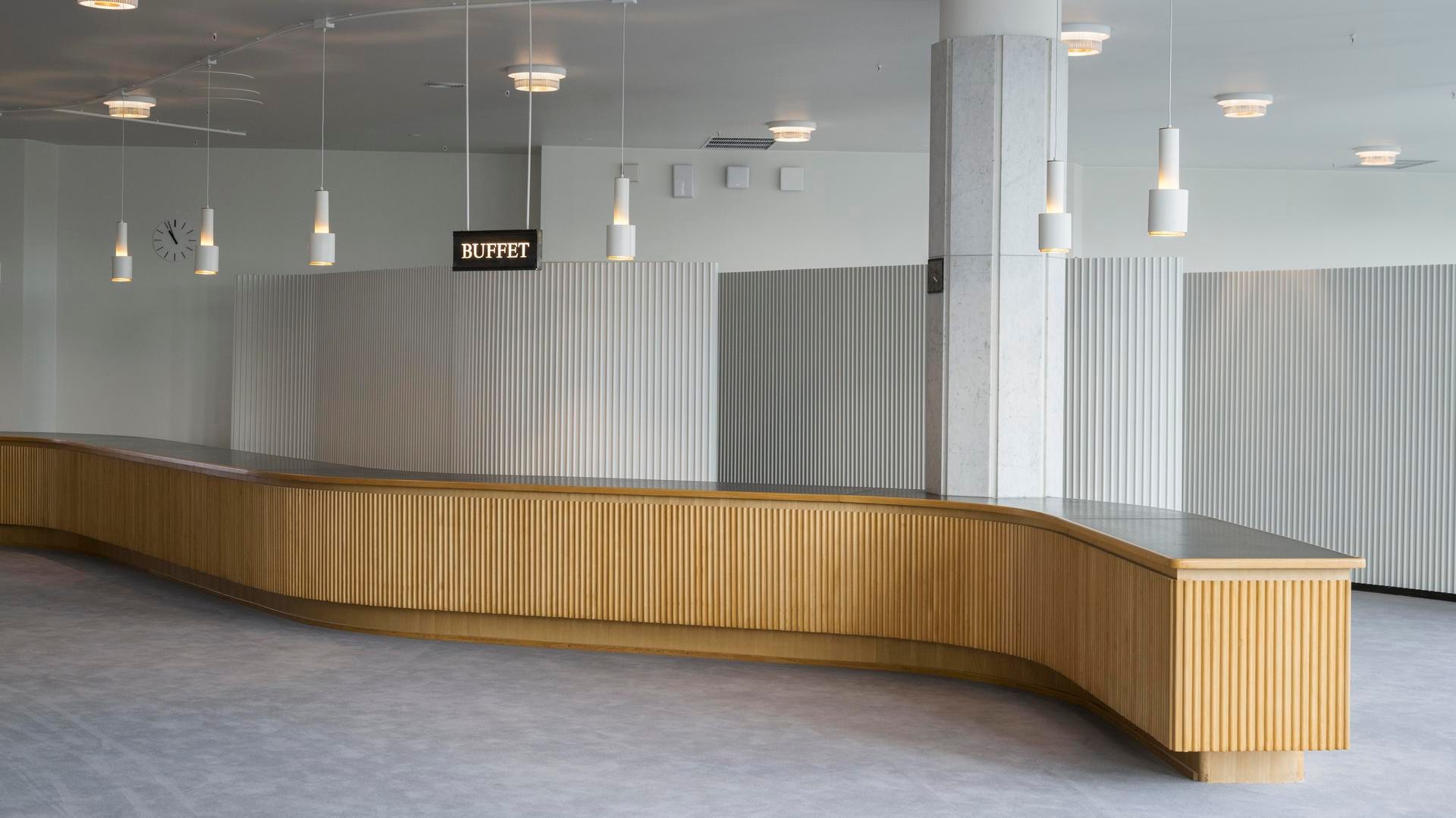 Moderni buffet-alue, jossa on kaareva puinen tiski ja riippuvat riippuvalaisimet tilavassa huoneessa.