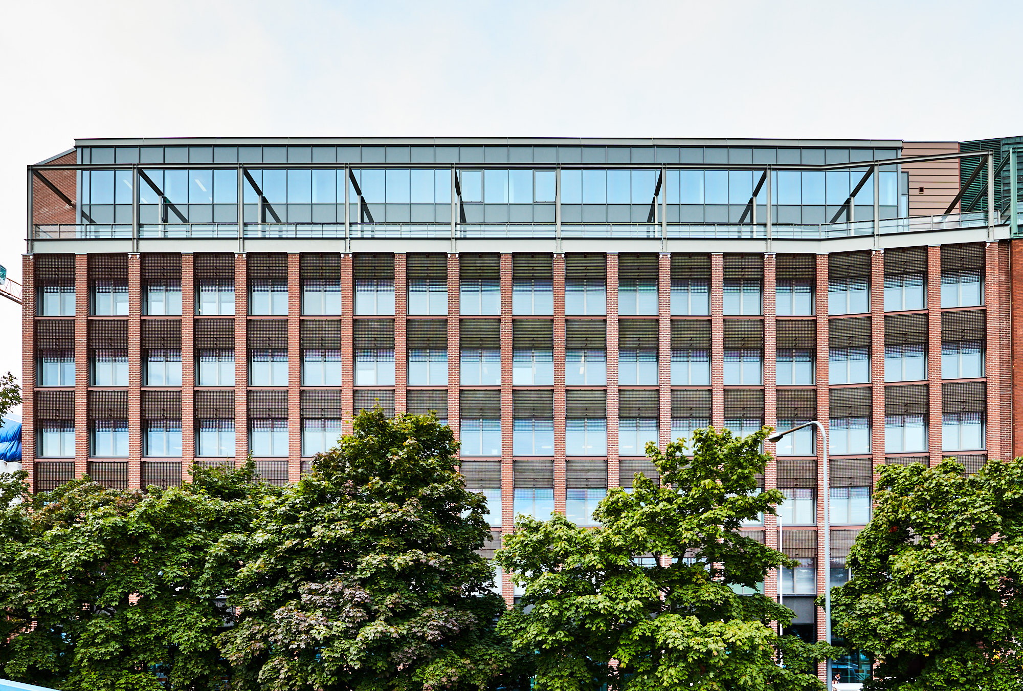 Tiilirakennus, jossa on suuret ikkunat, lasirakenteen peittämänä, vehreiden puiden ympäröimänä.