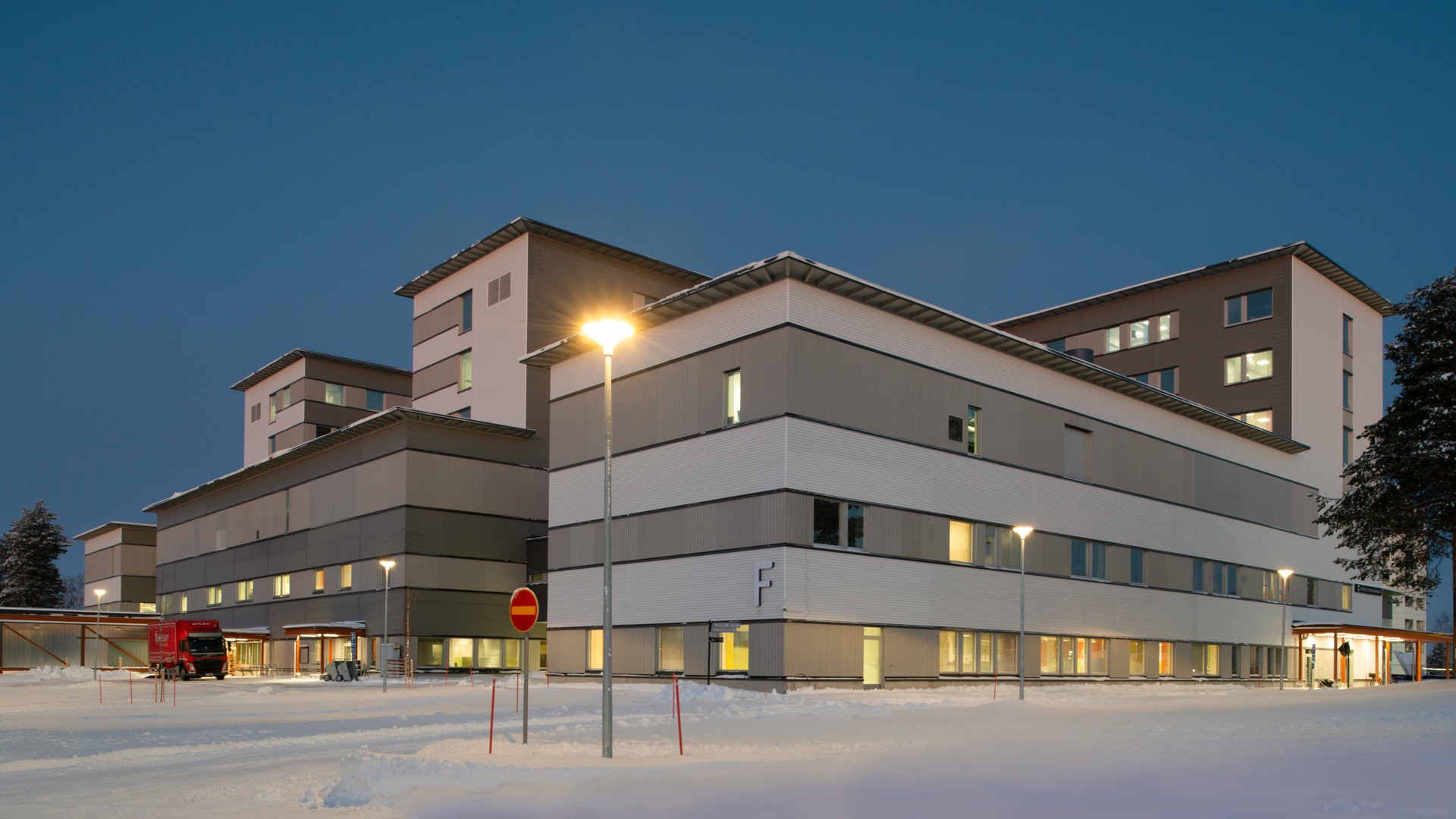 Kainuun uusi sairaala Moderni sairaalarakennus, jossa on valaistut ikkunat, ympäröitynä lumella hämärän taivaan alla.