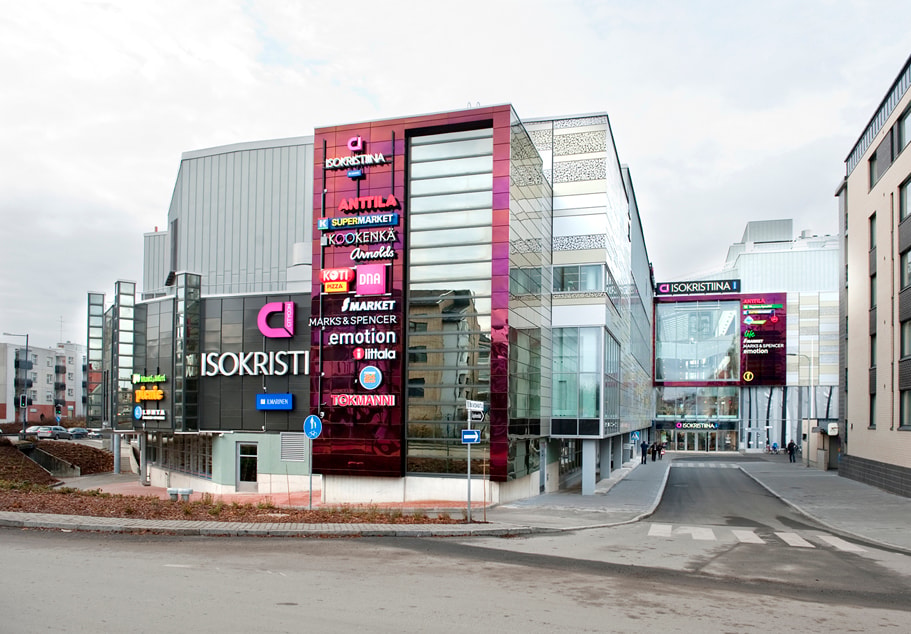 Moderni ostoskeskus lasijulkisivuilla ja erilaisilla myymäläkyltteillä urbaanissa ympäristössä.