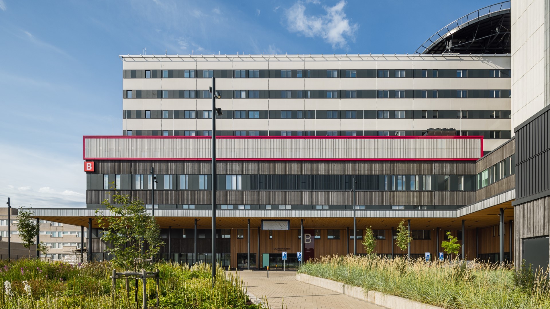 Moderni sairaalarakennus, jossa on punareunainen osio, merkitty kirjaimella 'B', jota ympäröi maisemoitu vihreys.