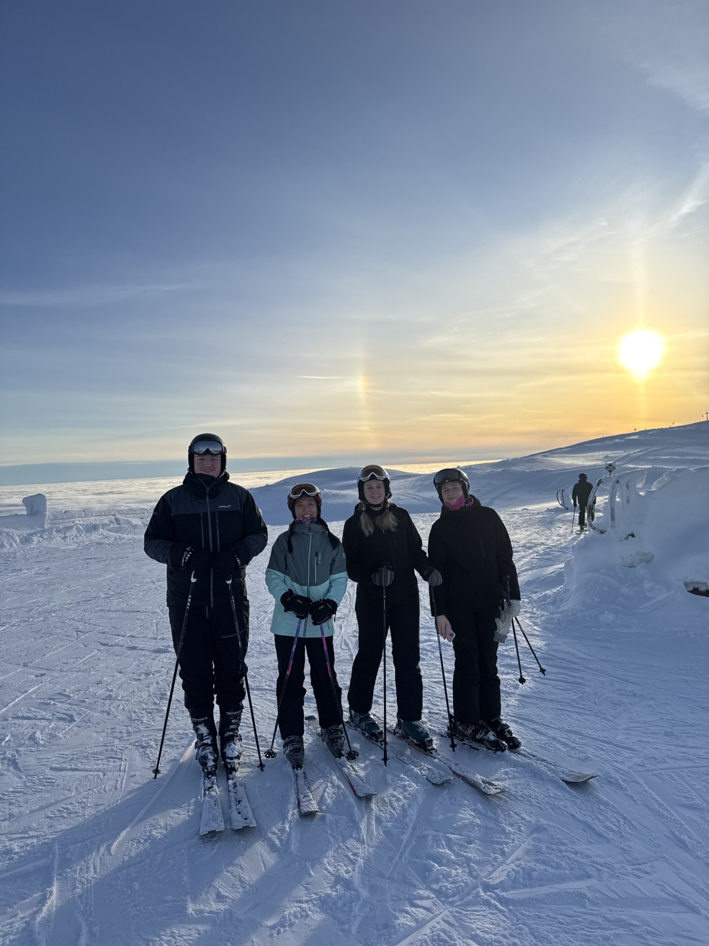 Fire skiløpere står på en snødekt skråning ved solnedgang, utstyrt med ski og staver, og nyter vinteridrett.