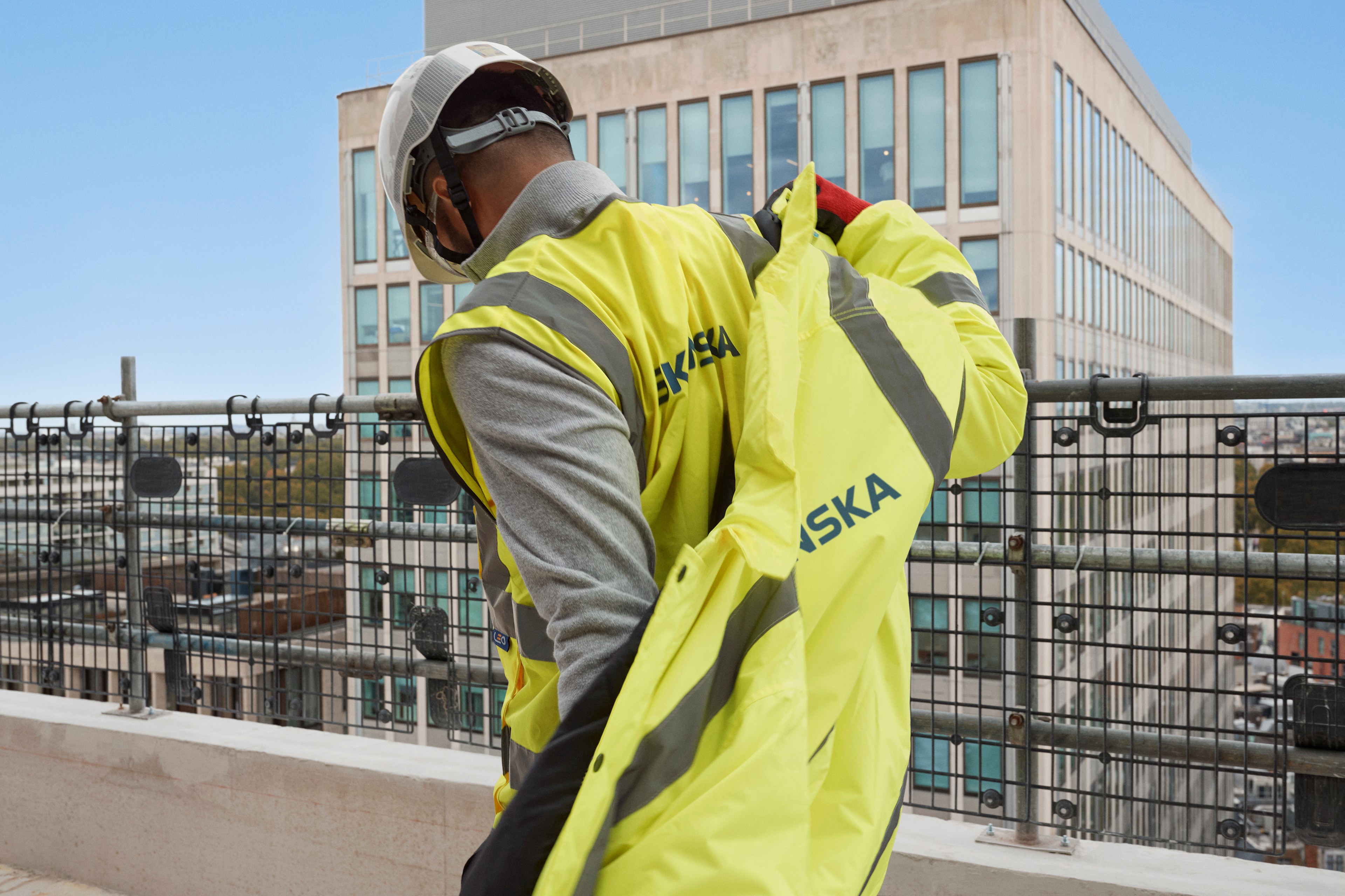 Construction Worker in Safety Gear on a Building Site Construction worker in Skanska safety gear adjusts vest on rooftop with city buildings in background.