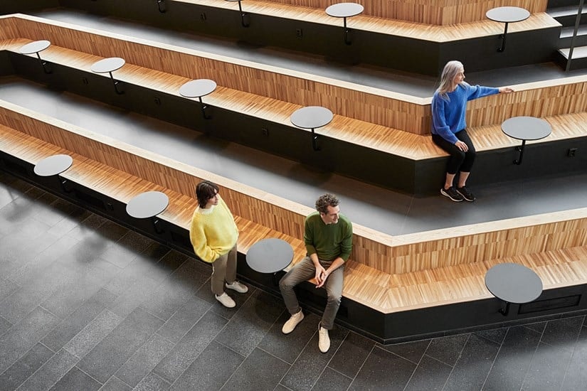 Moderní aula s patrovými dřevěnými sedadly, malými kulatými stolky a třemi osobami sedícími samostatně.