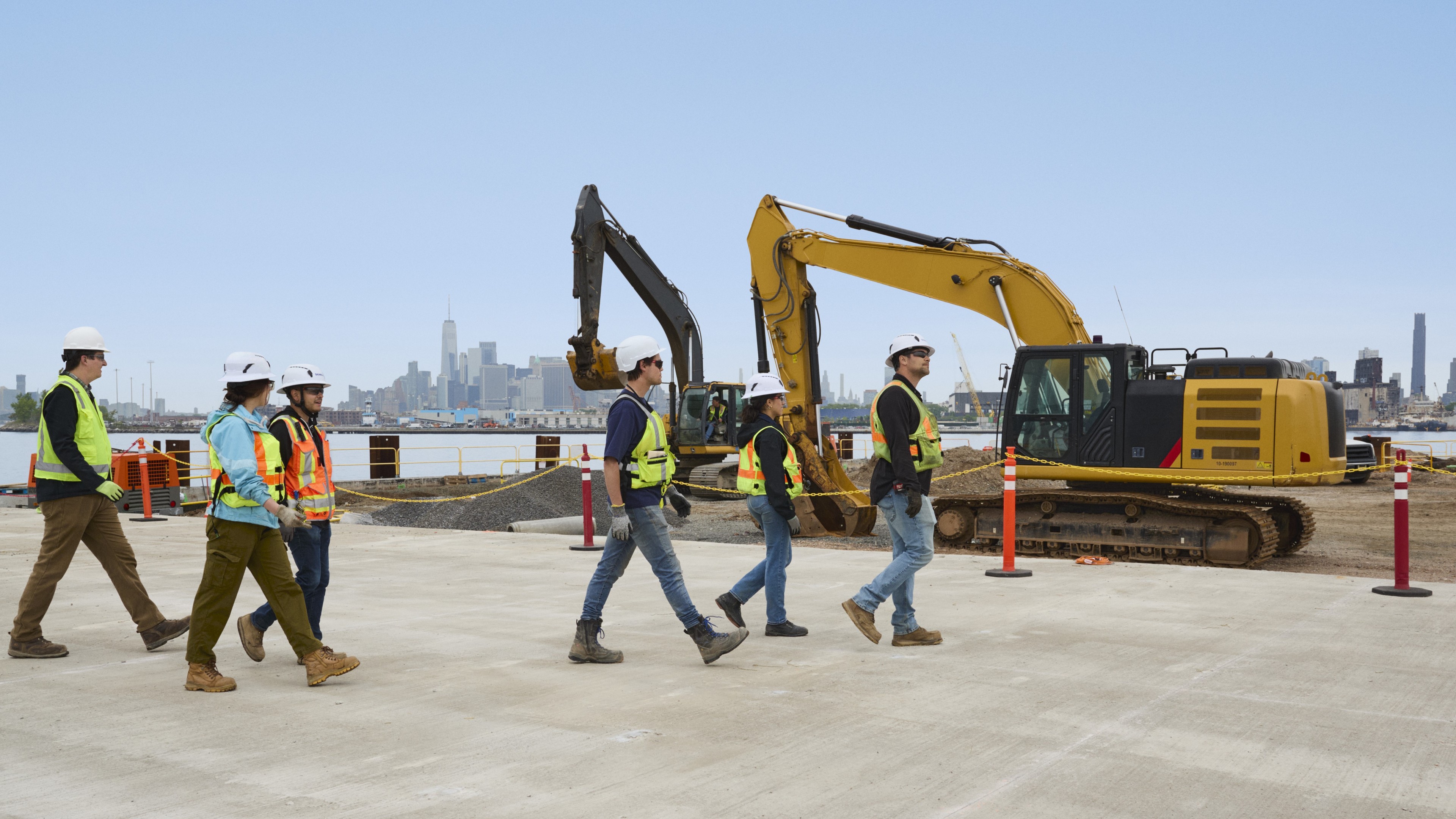Construction workers in safety gear walk on a site with excavators and city skyline in the background.