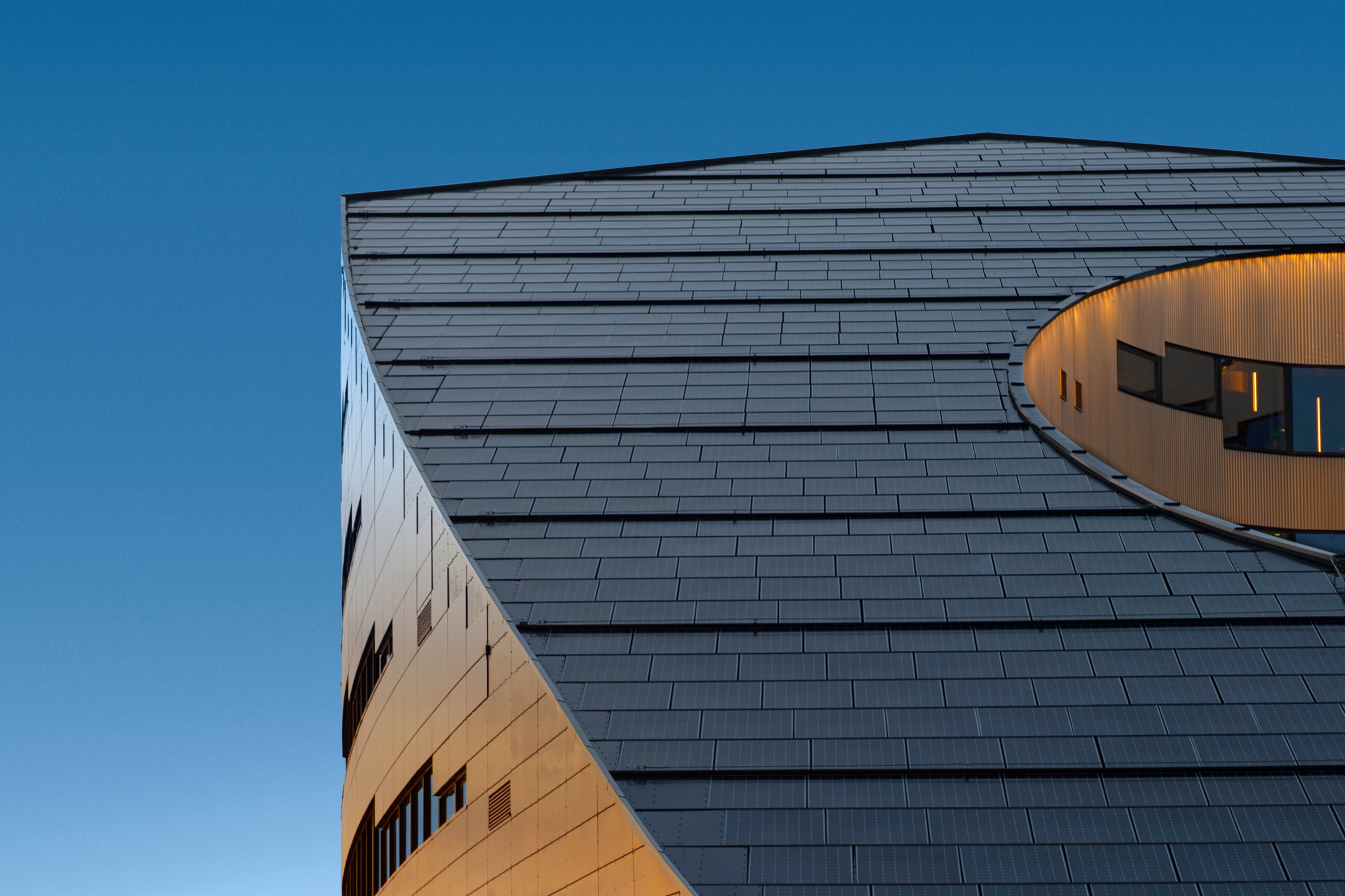 Modern angular building with a circular cutout, featuring sleek metal panels against a blue sky.