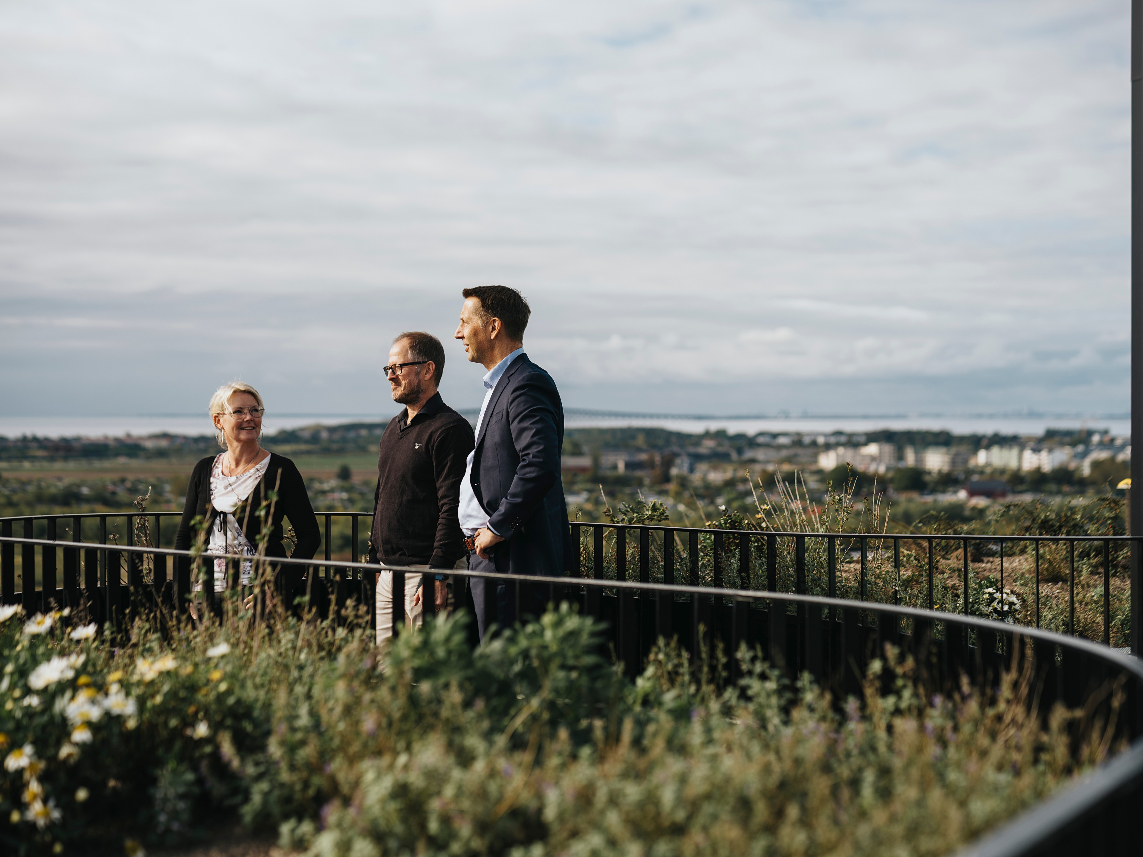 Tre personer står vid ett räcke och blickar ut över ett naturskönt landskap med molnig himmel.