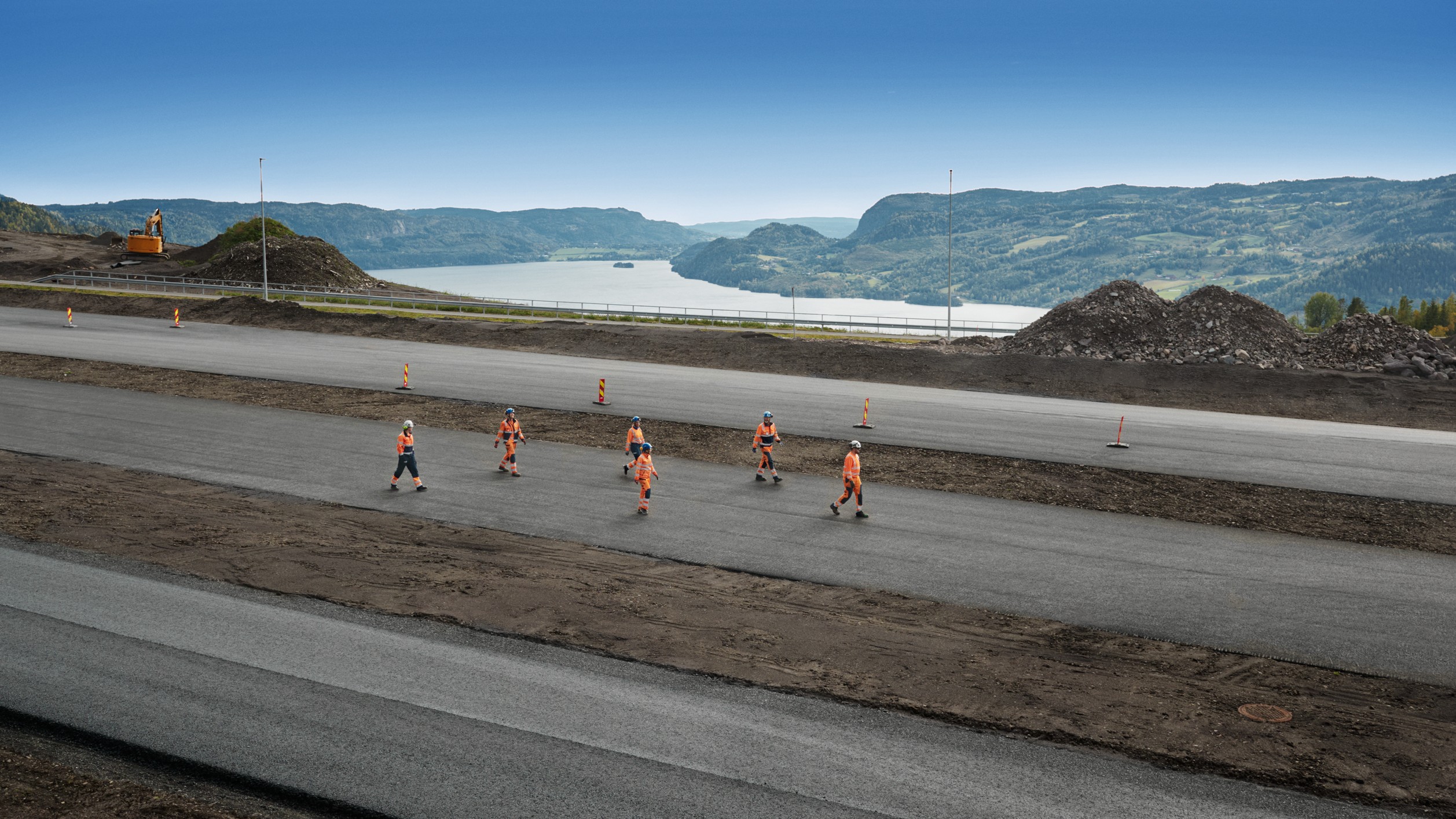Construction workers in orange vests walk on a newly paved road with a scenic lake view.