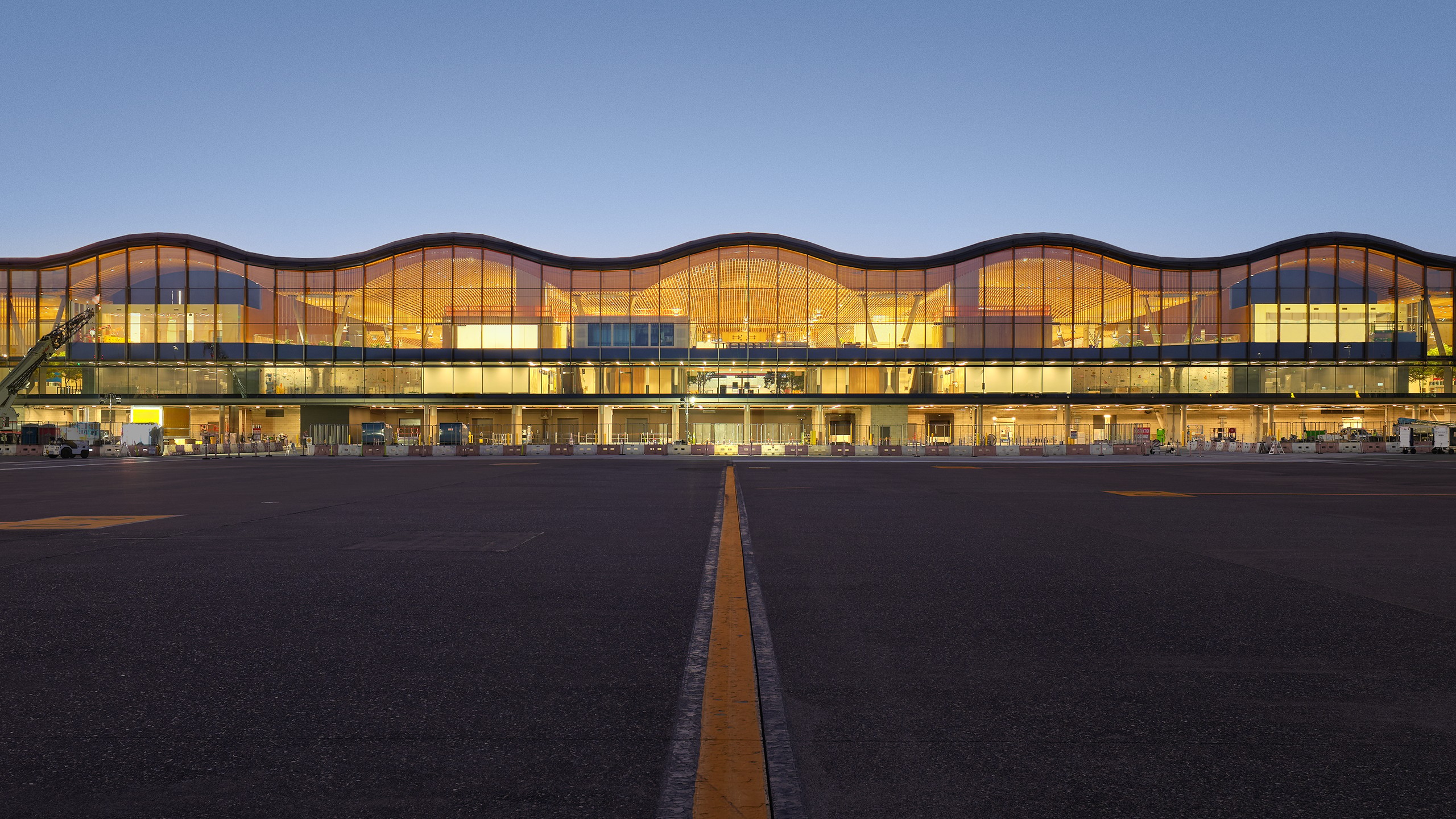 Modern airport terminal with wavy roof design and illuminated glass facade at dusk.