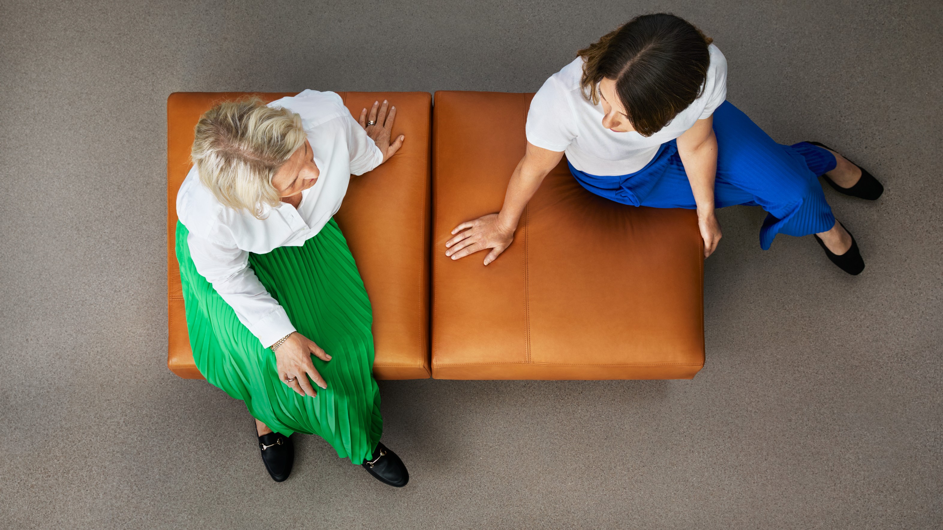 Two Women Sitting and Conversing in Stylish Outfits Two women in colorful skirts converse while seated on a modern, brown leather bench.