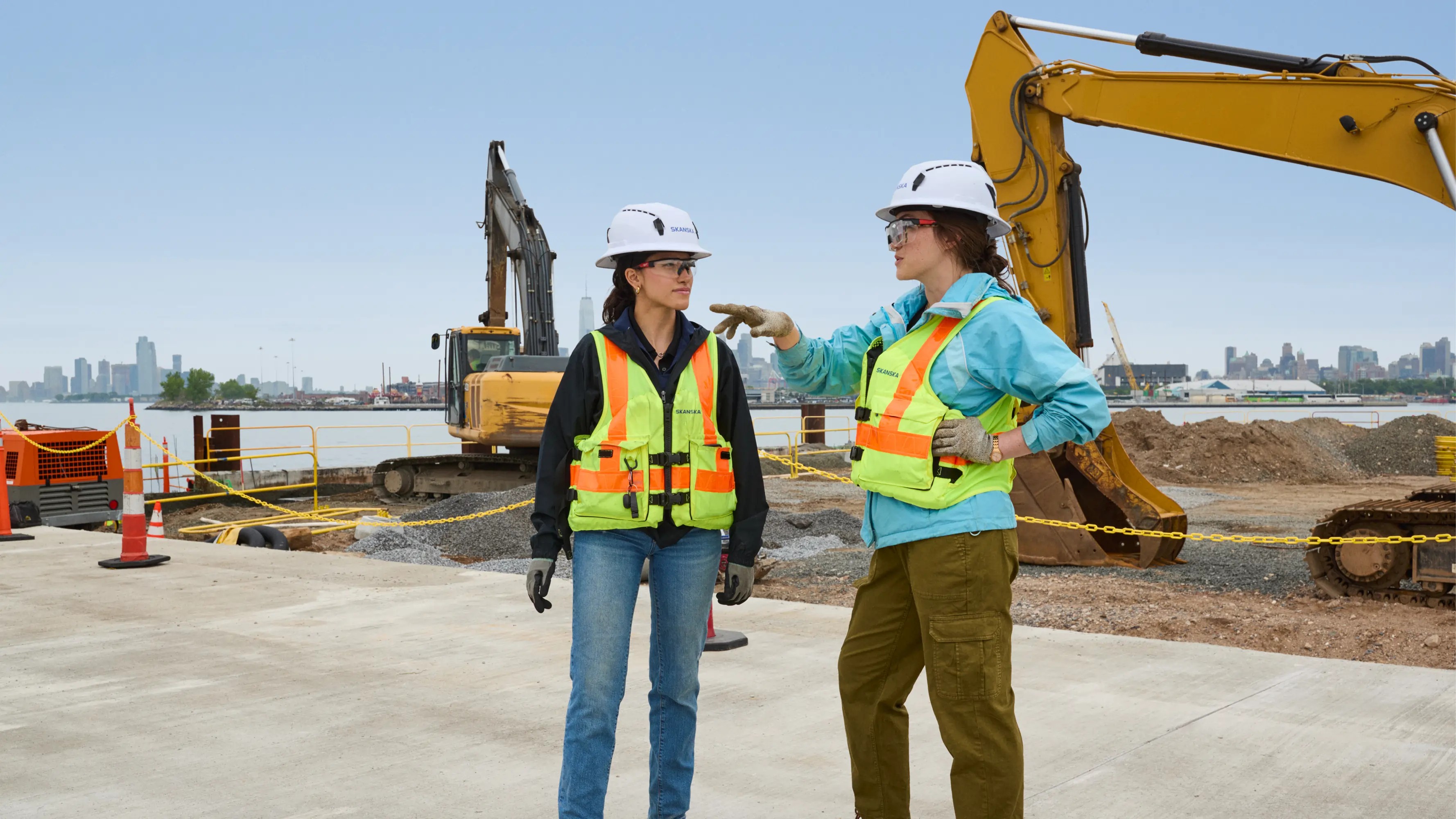 Two construction workers wearing safety gear converse at an active construction site.