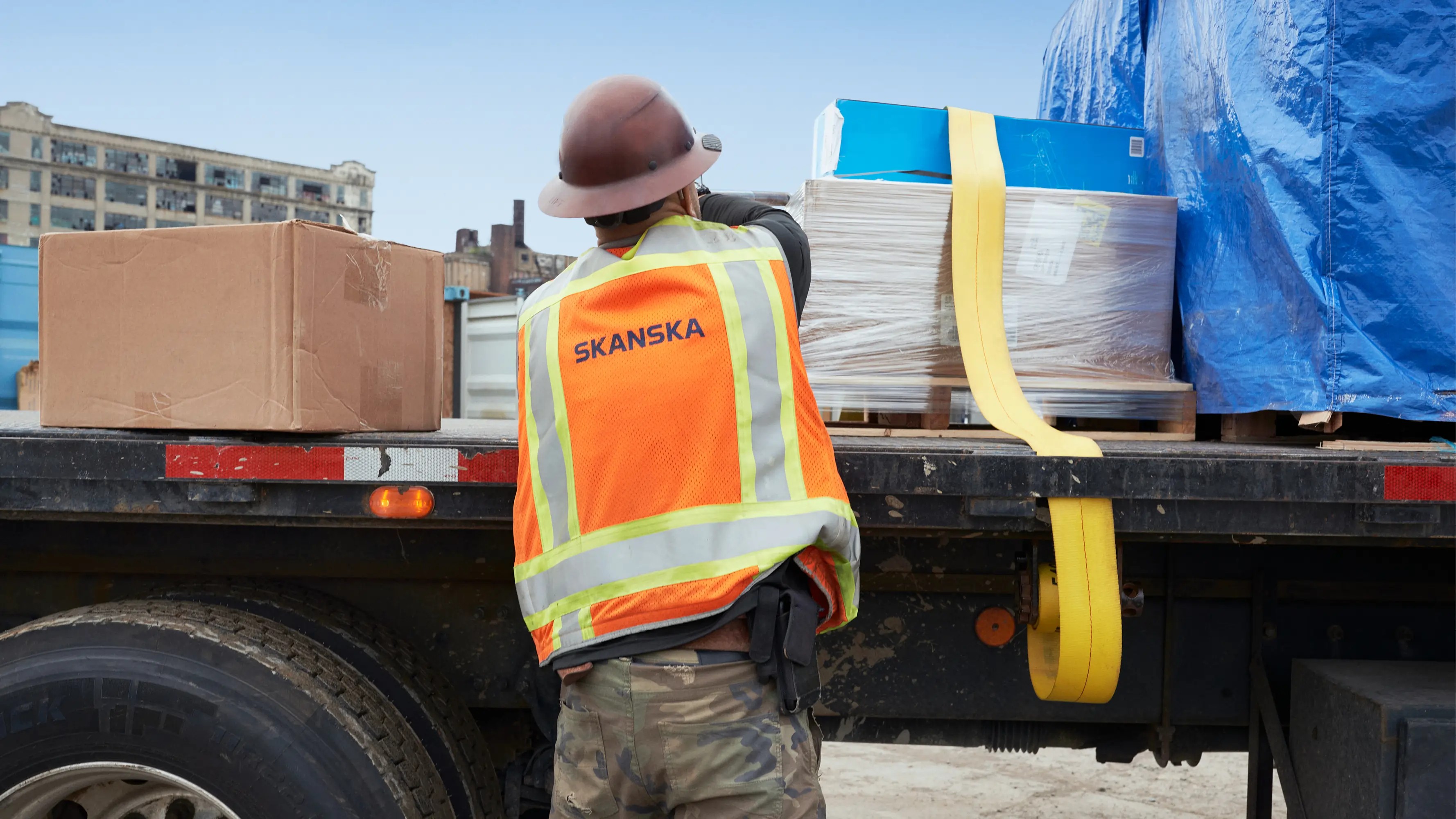 Construction worker in Skanska safety vest secures cargo on a flatbed truck with straps.