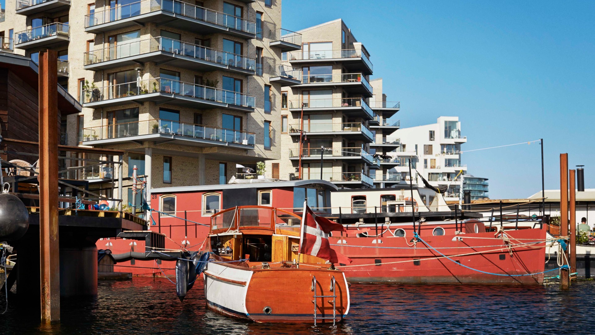 Red wooden boat with Danish flag docked near modern apartment buildings under clear sky.